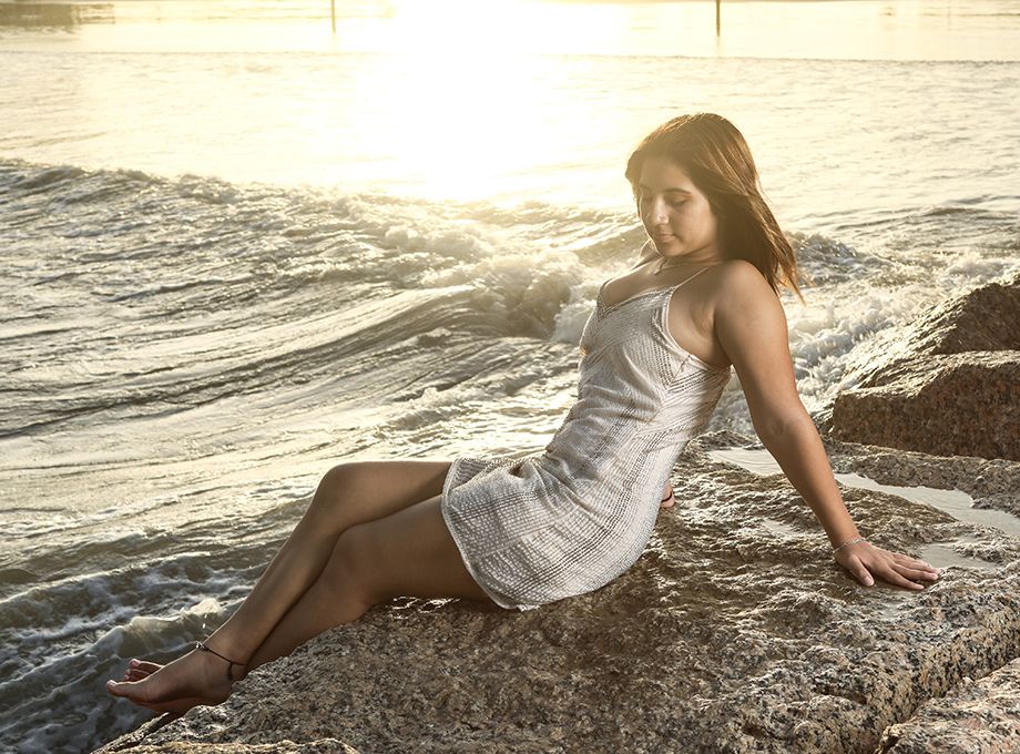Model in white sundress poses on rocky beach at sunset.
