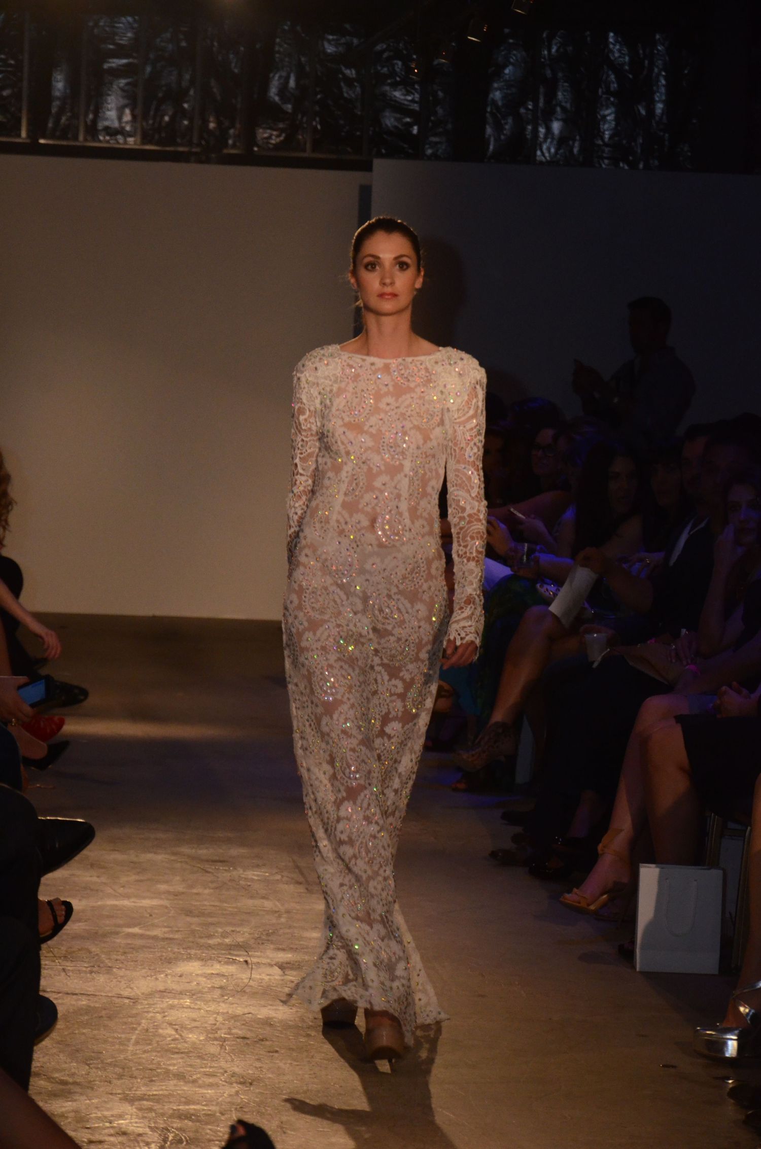 Model walks runway wearing elegant floor-length beaded lace gown during fashion show.