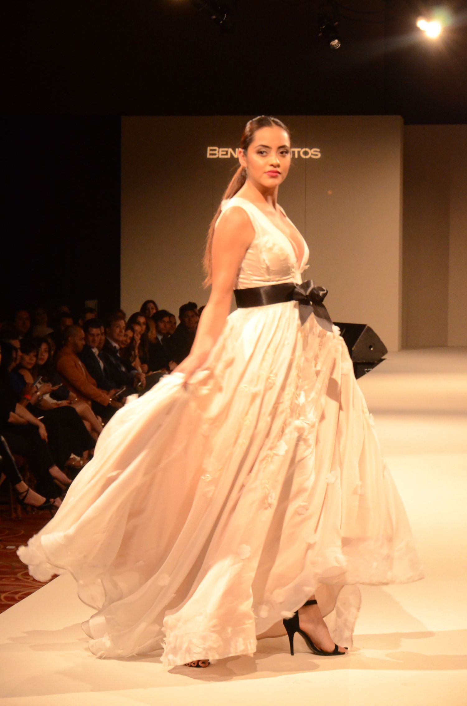 Model walks runway in flowing white ballgown with black sash at fashion show.
