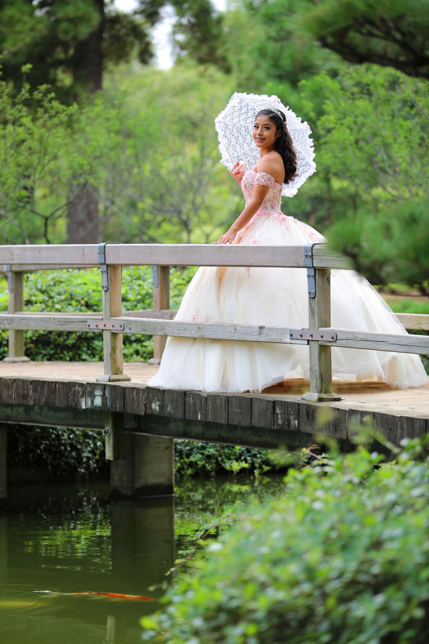 Romantic portrait with white parasol and gown on wooden bridge over pond.