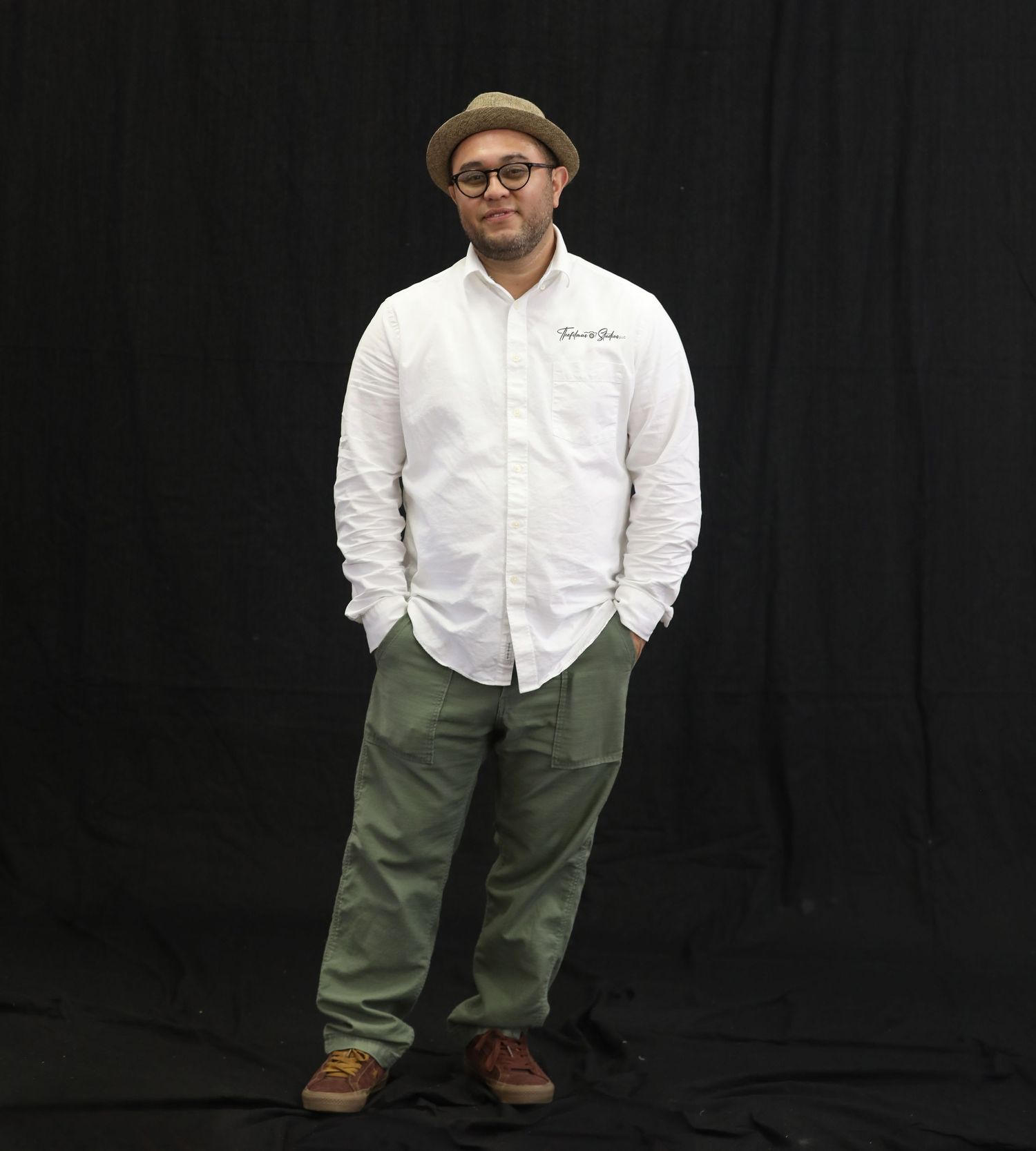 Person in casual white shirt and olive pants stands against black backdrop.