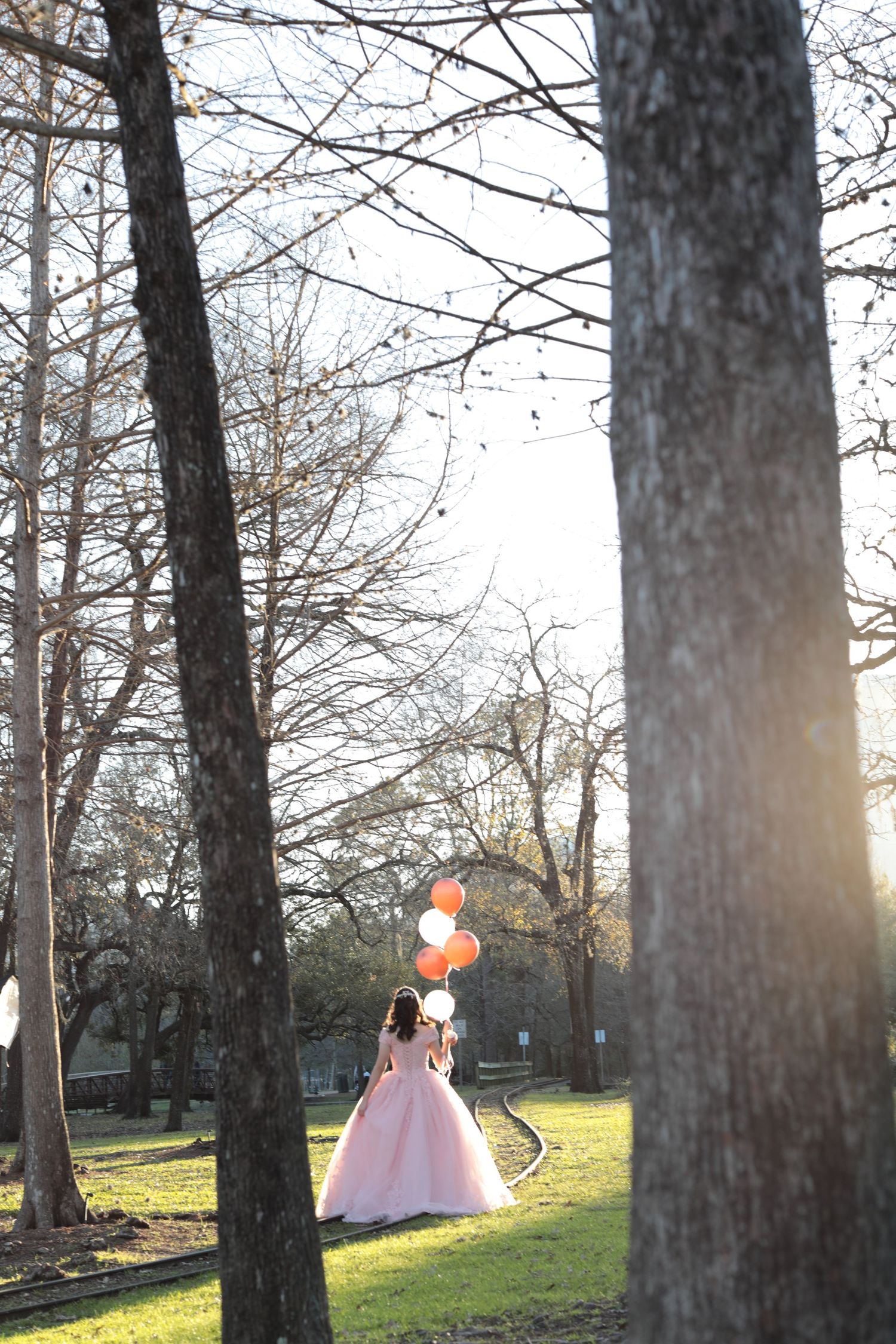 A person in a pink ball gown poses with orange balloons in a sunlit park setting.