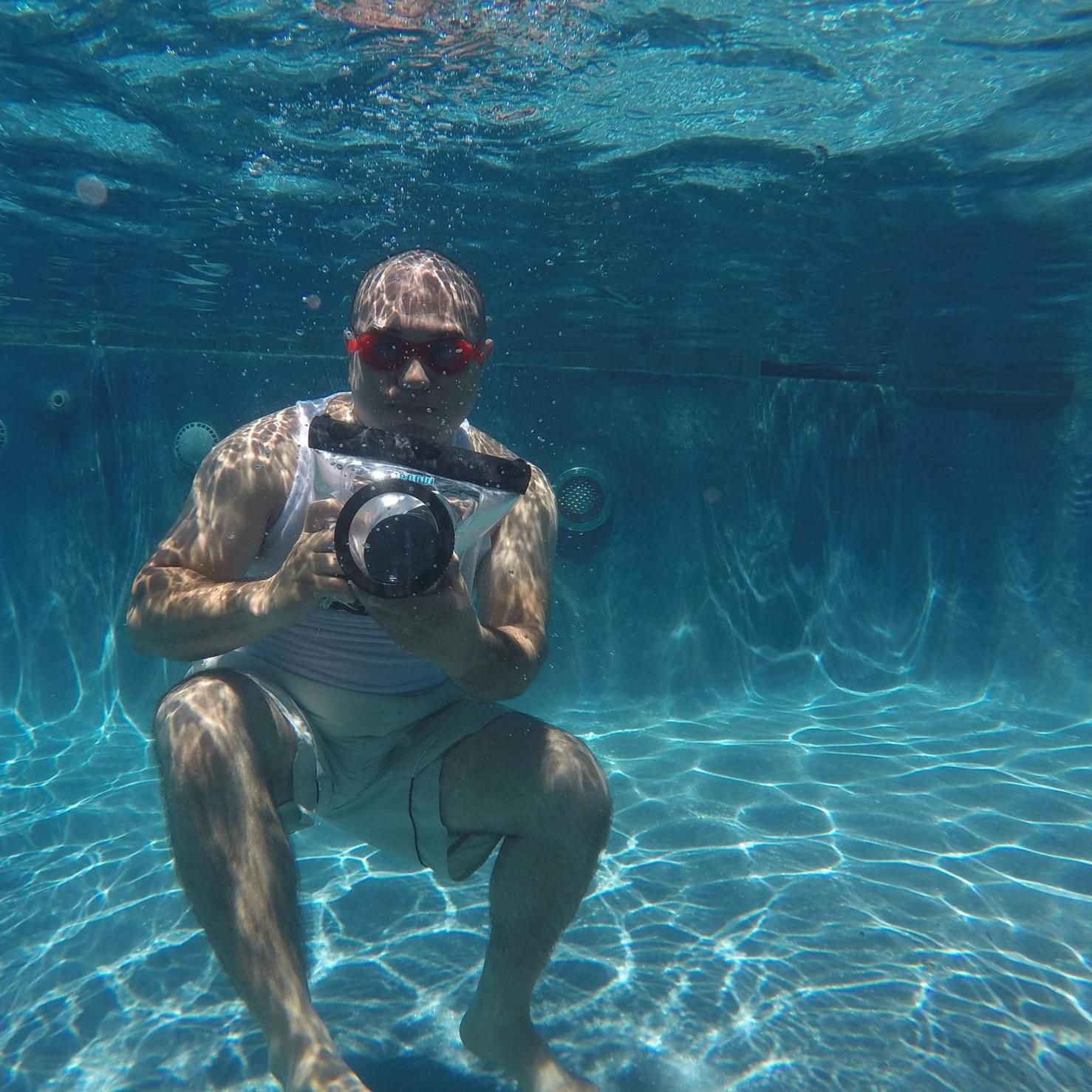 Underwater photographer with a digital camera sitting on pool floor with sunlight filtering through turquoise water.