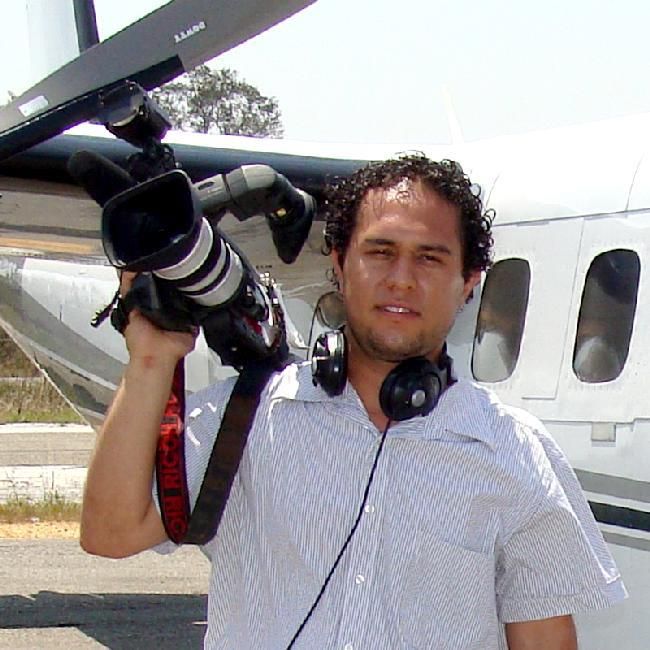 Professional photographer with camera equipment and headphones standing next to a small aircraft on a sunny day.