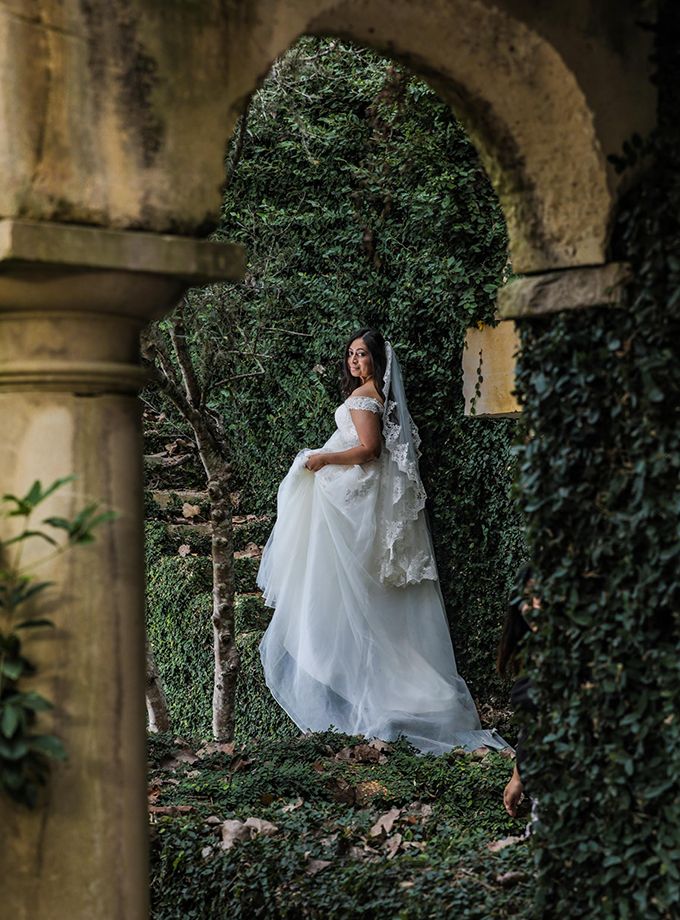 Bride in flowing white gown poses under stone archway surrounded by ivy.