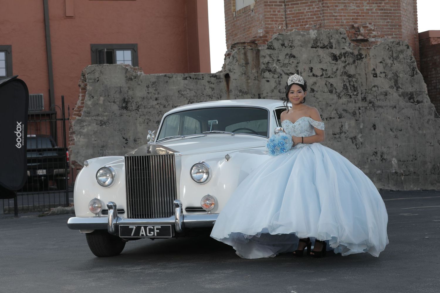 Elegant light blue ballgown posed next to vintage white Rolls Royce against stone wall.