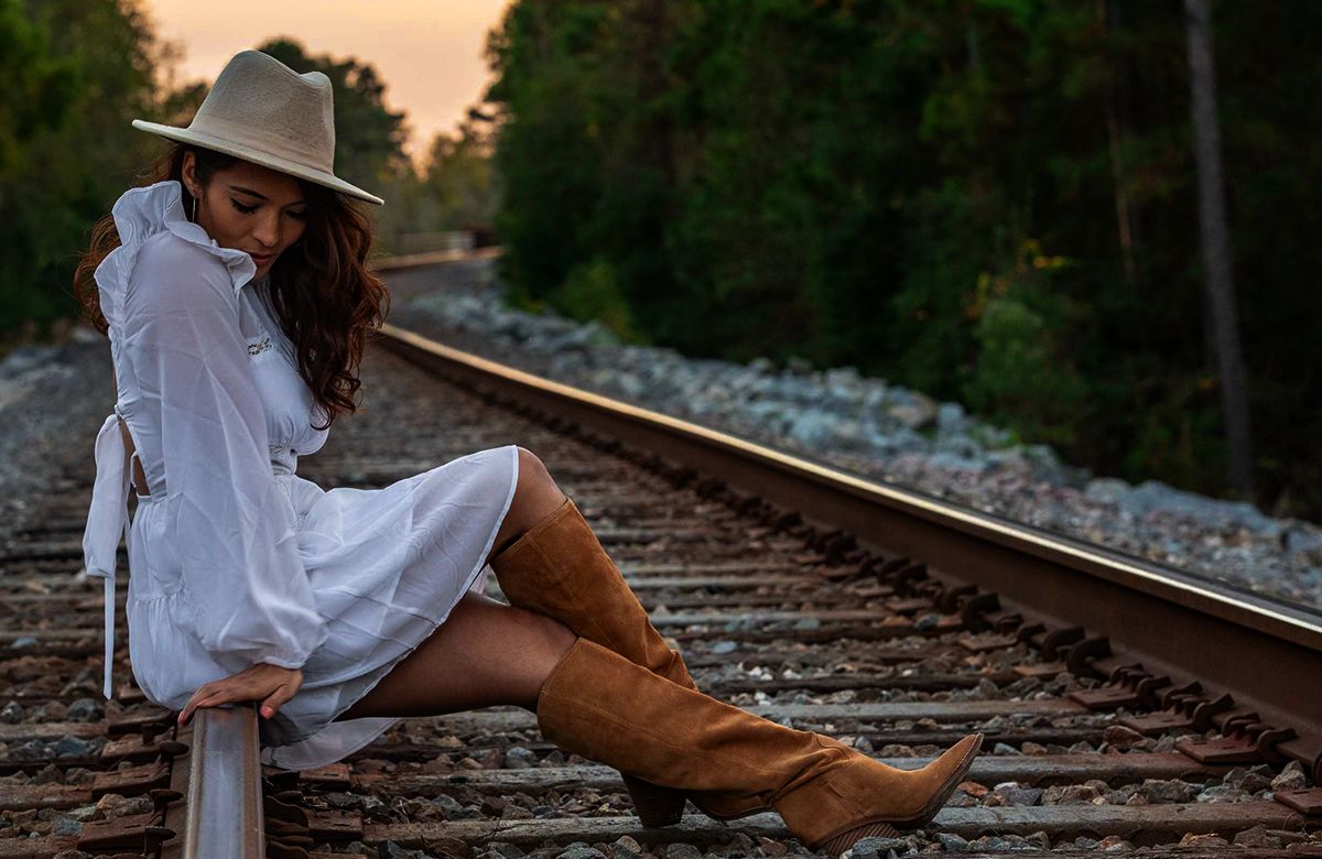 Atmospheric photo of person in white dress and cowboy boots sitting on railroad tracks at sunset.