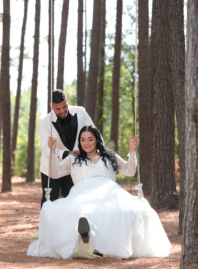 A couple in wedding attire playfully interacts on a tree-lined path with a white dress flowing.