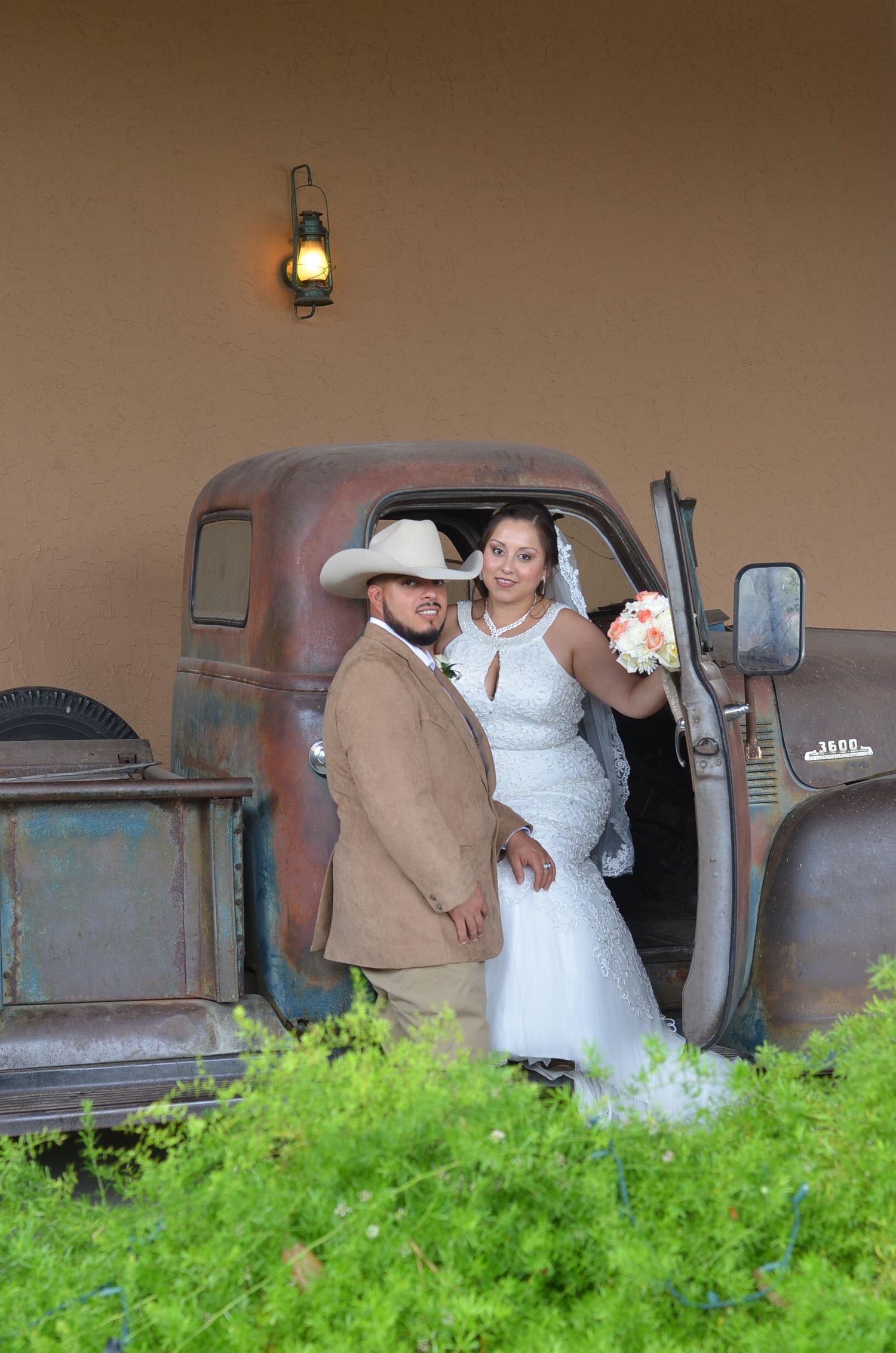 A couple poses by a vintage truck in wedding attire with rustic wall and lantern backdrop.