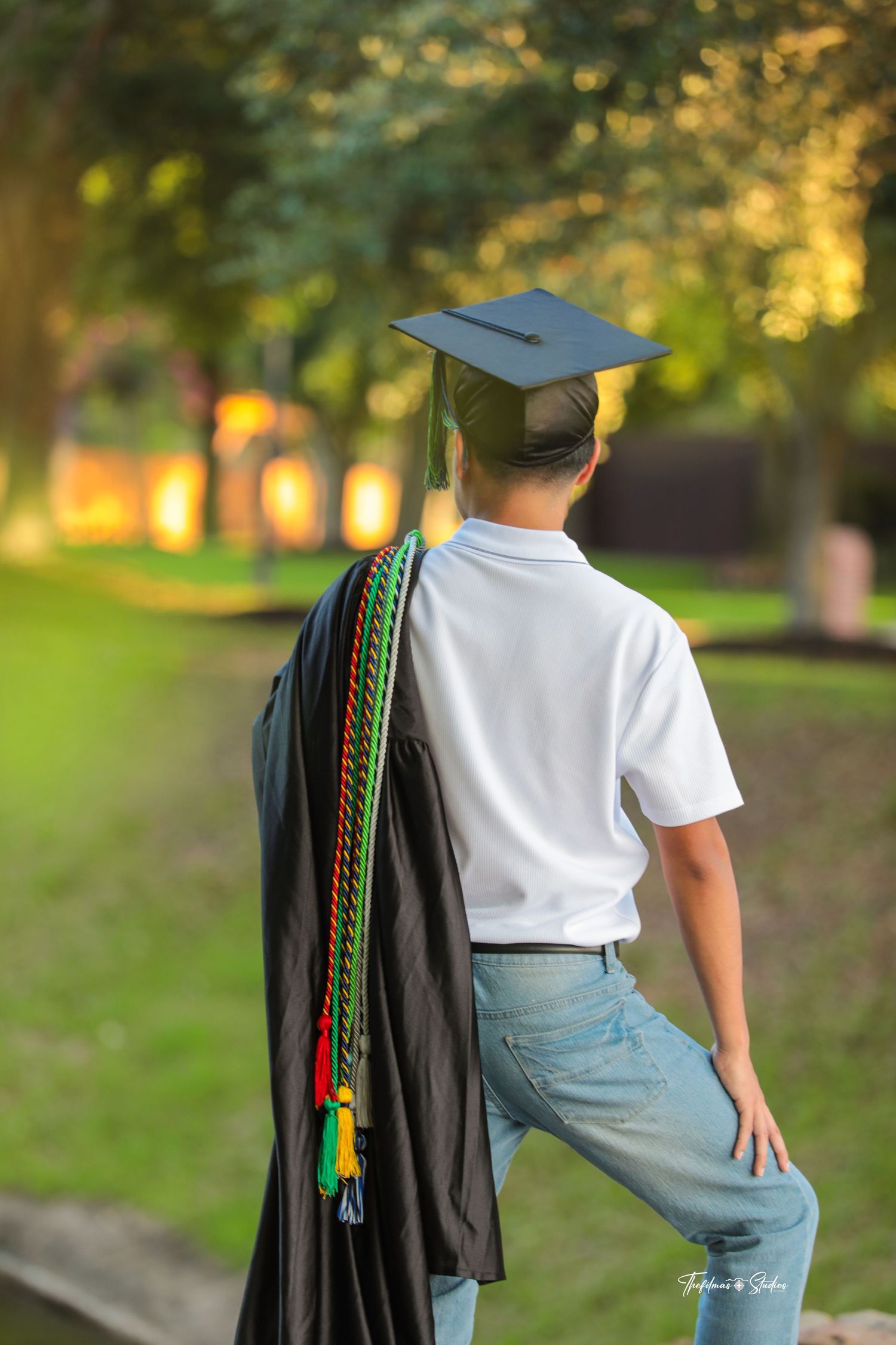Graduate wearing cap and gown with colorful cords poses on campus path.