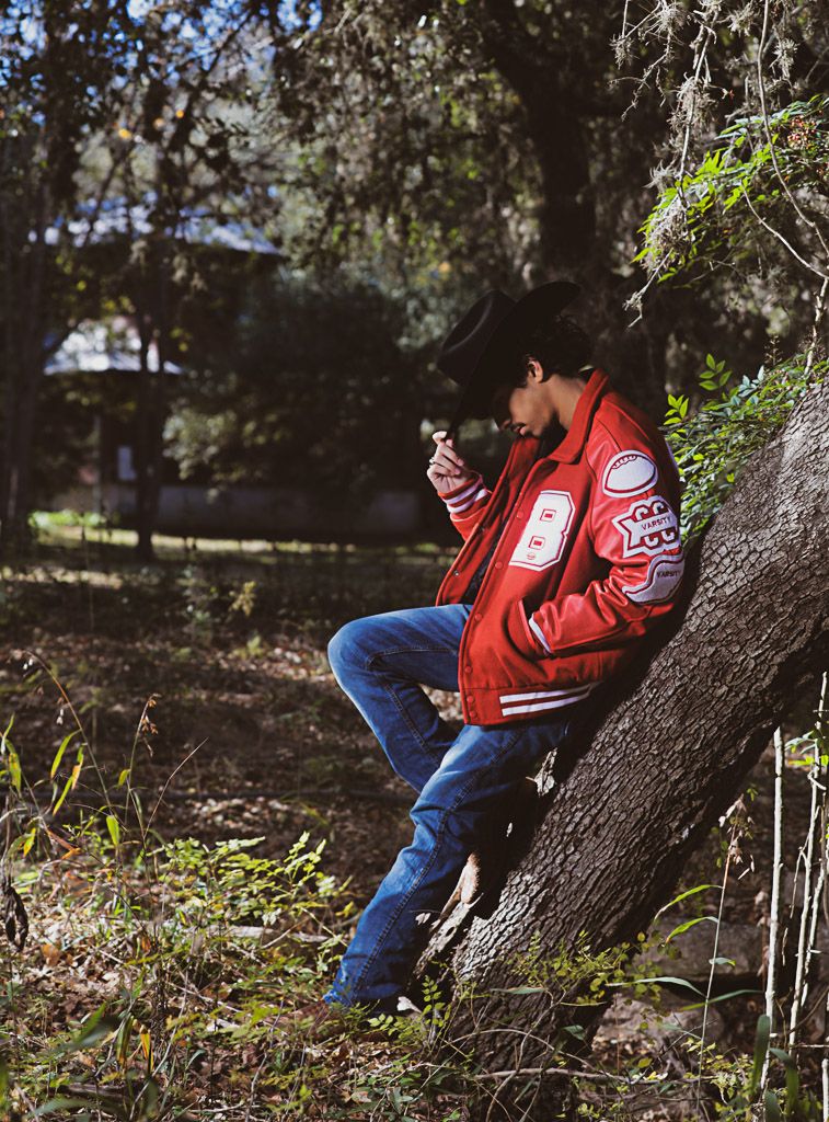 Person in red varsity jacket and jeans sitting on tree trunk in wooded setting.