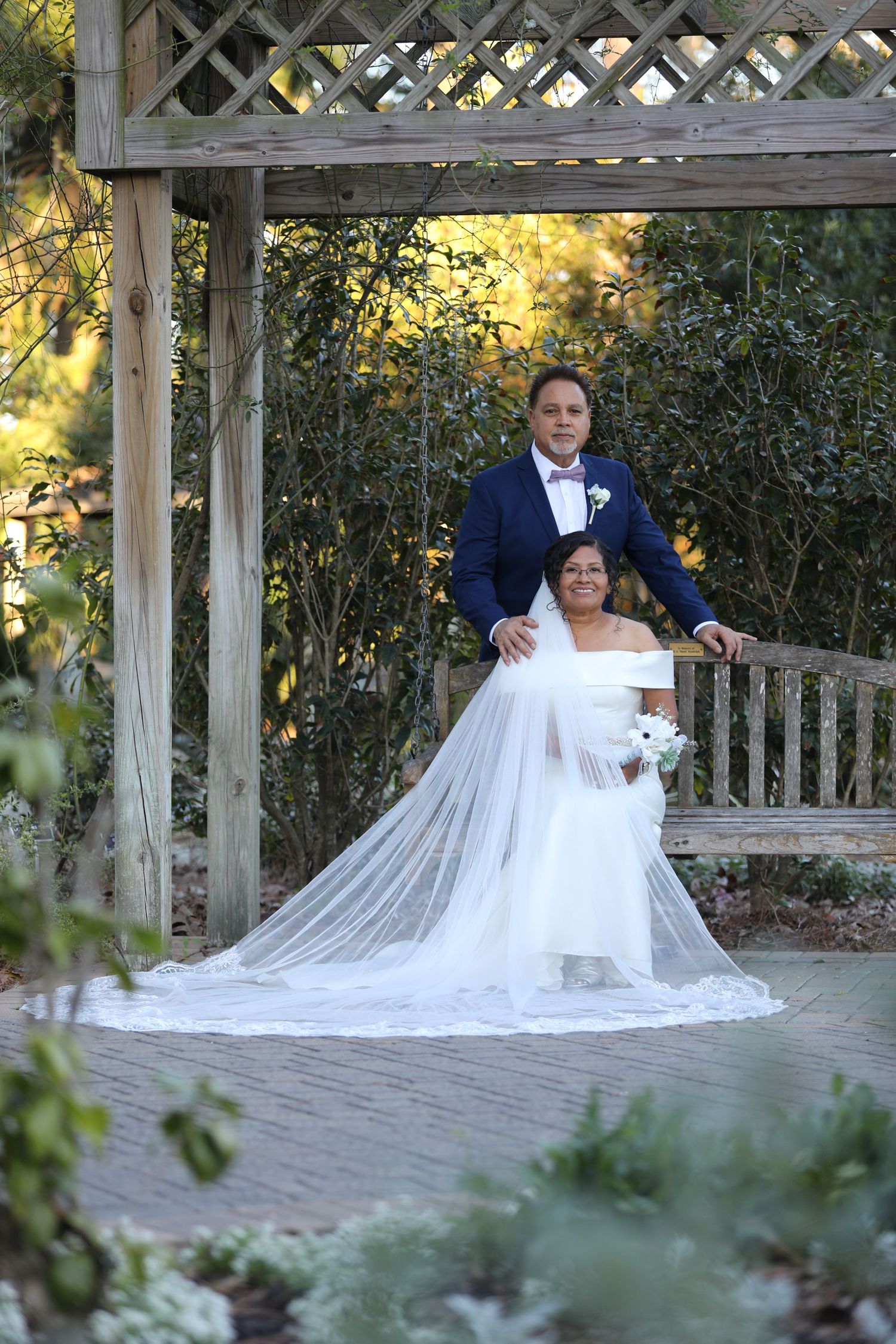 Wedding couple posing under wooden garden arbor surrounded by greenery.