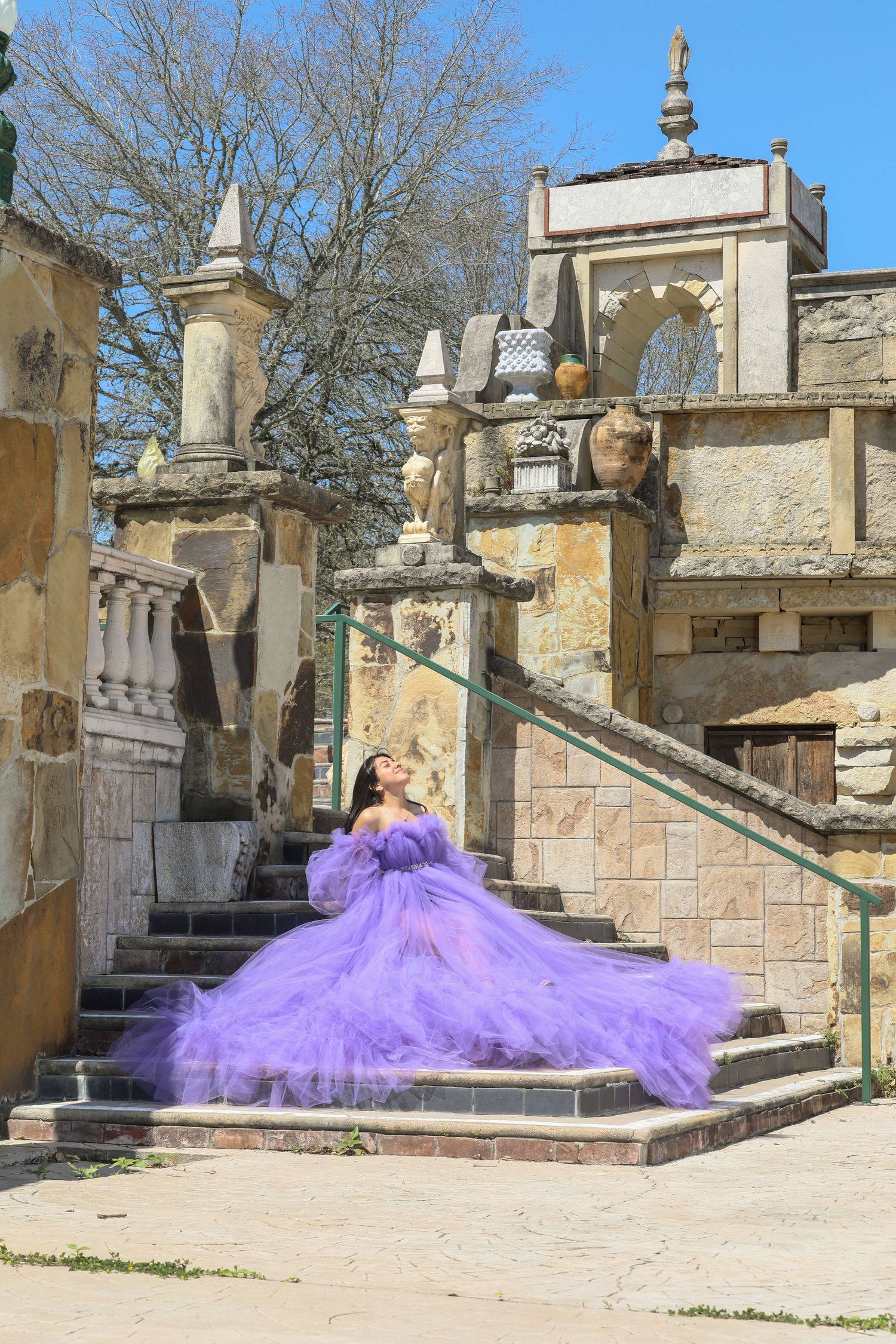 A flowing purple tulle ball gown cascades down ornate stone steps of a classical architectural setting.
