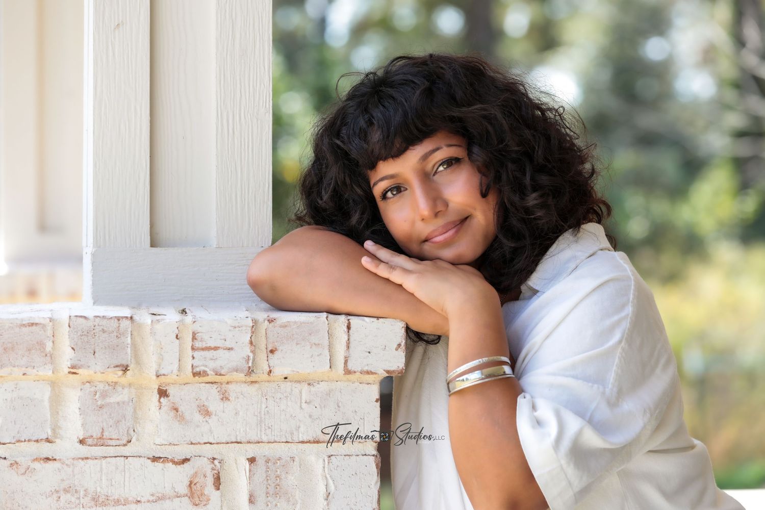 A woman in a white top leans against a white column while posing outdoors in natural lighting.