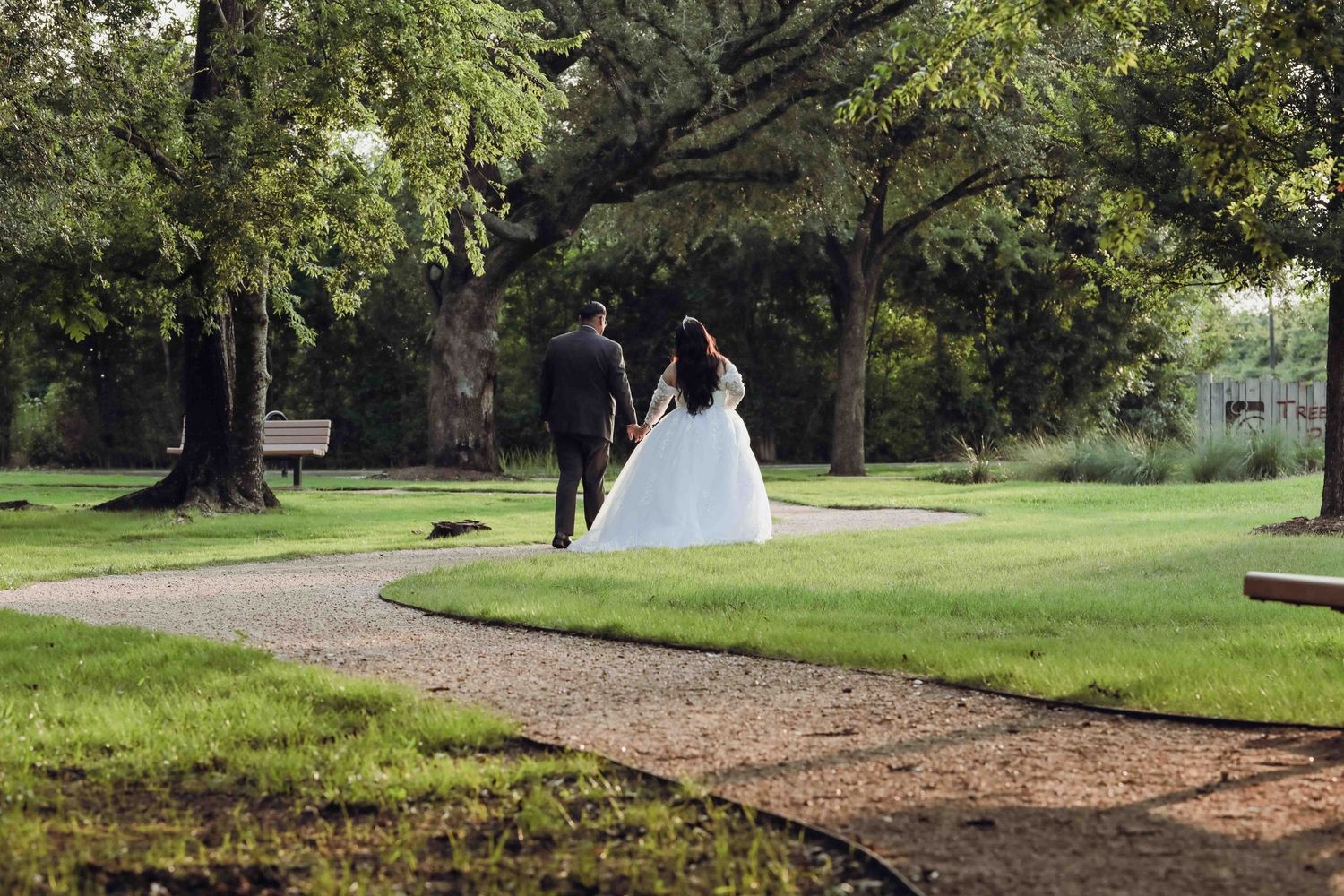 Two newlyweds walk along a gravel path through a lush green garden setting on their wedding day.