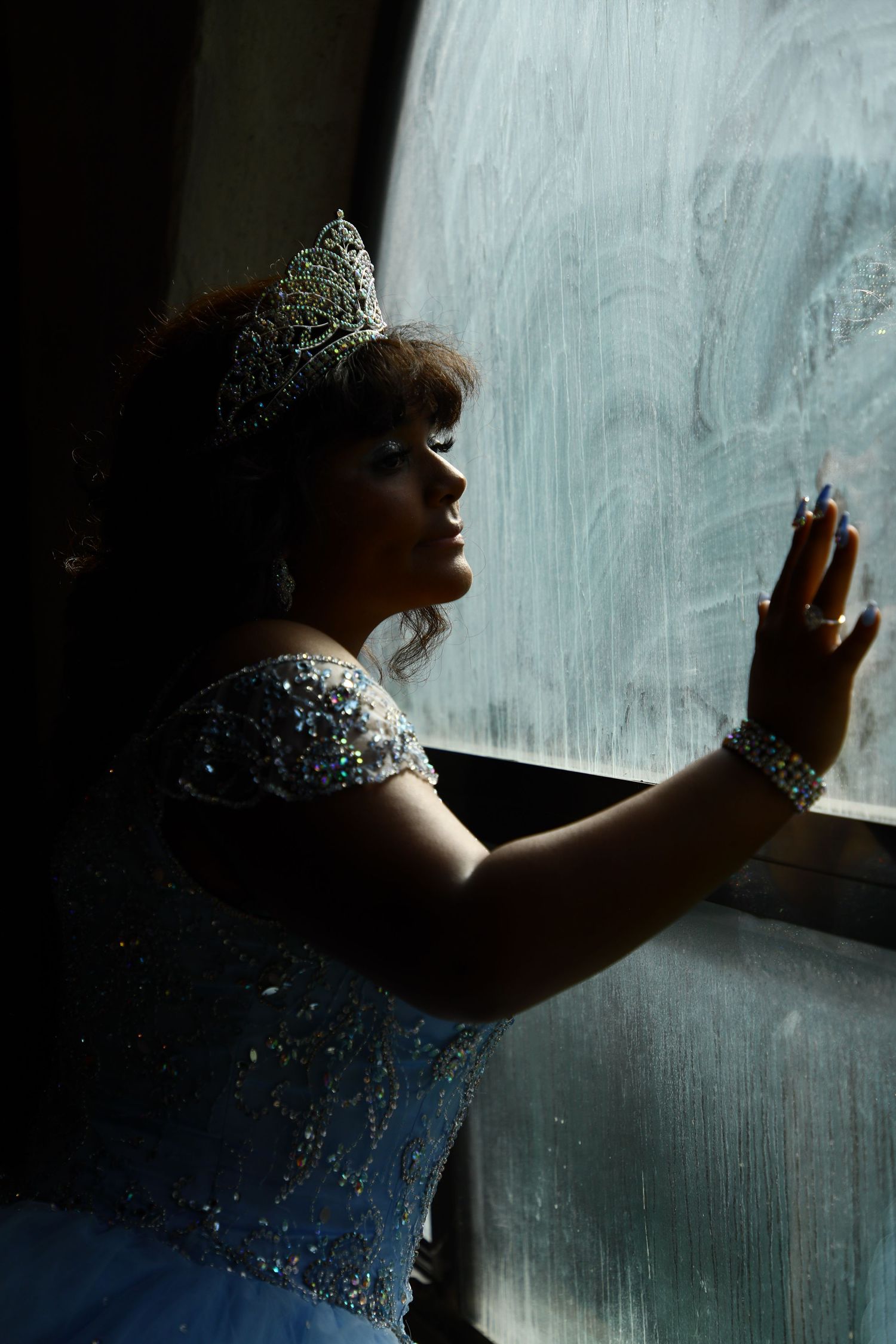 A silhouette in a sparkly blue gown and tiara gazes out a window in dramatic lighting.