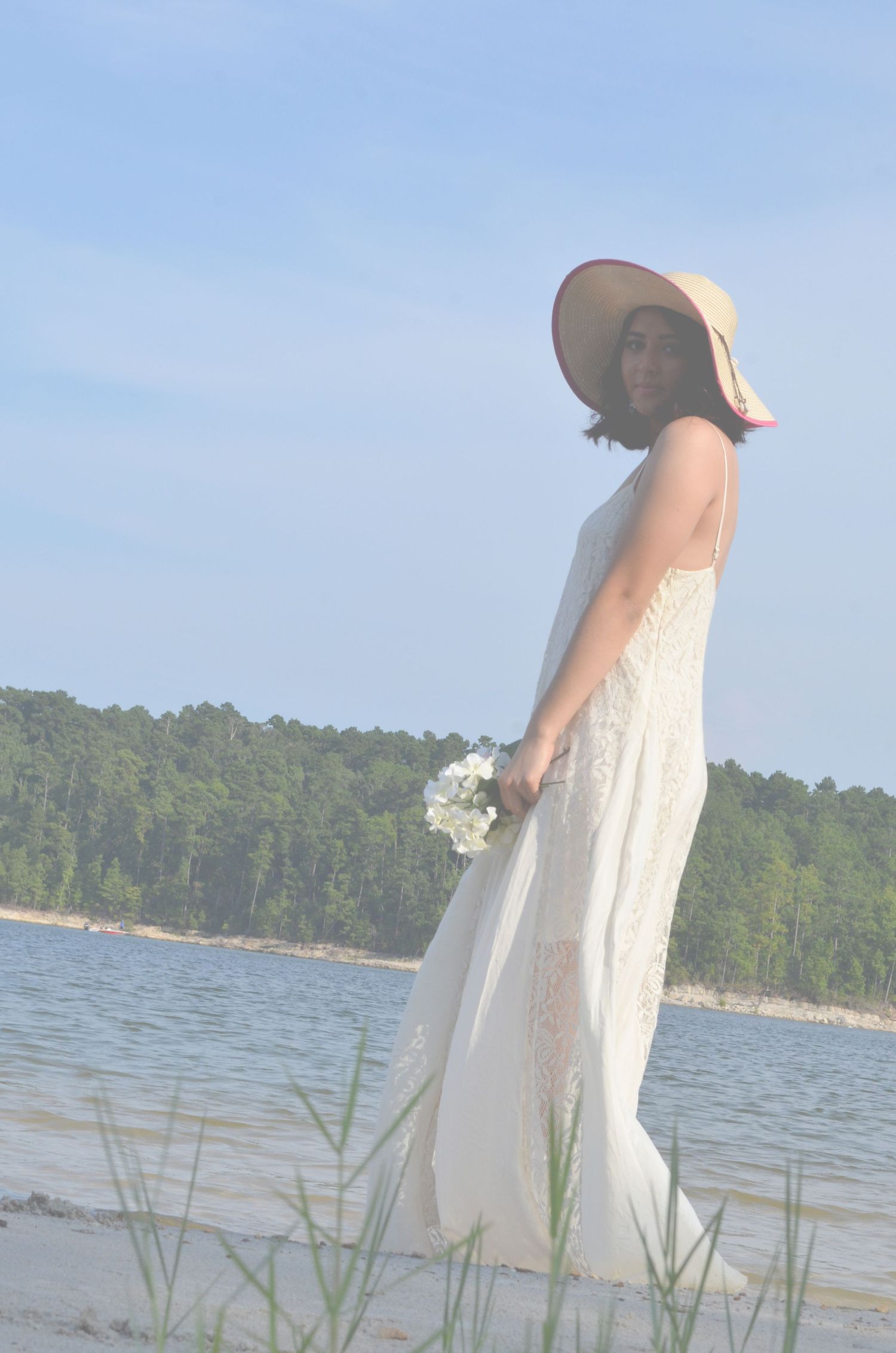 Figure in white dress and sunhat stands by lakeside holding bouquet.