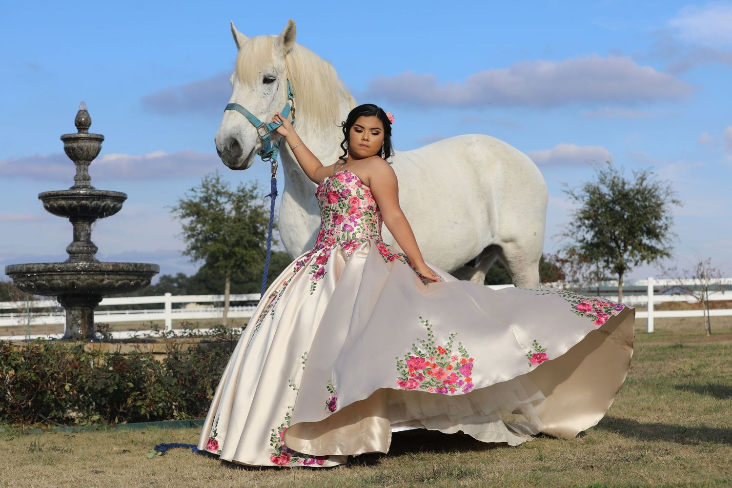 Elegant dress with floral embellishments photographed with white horse and fountain in countryside setting.
