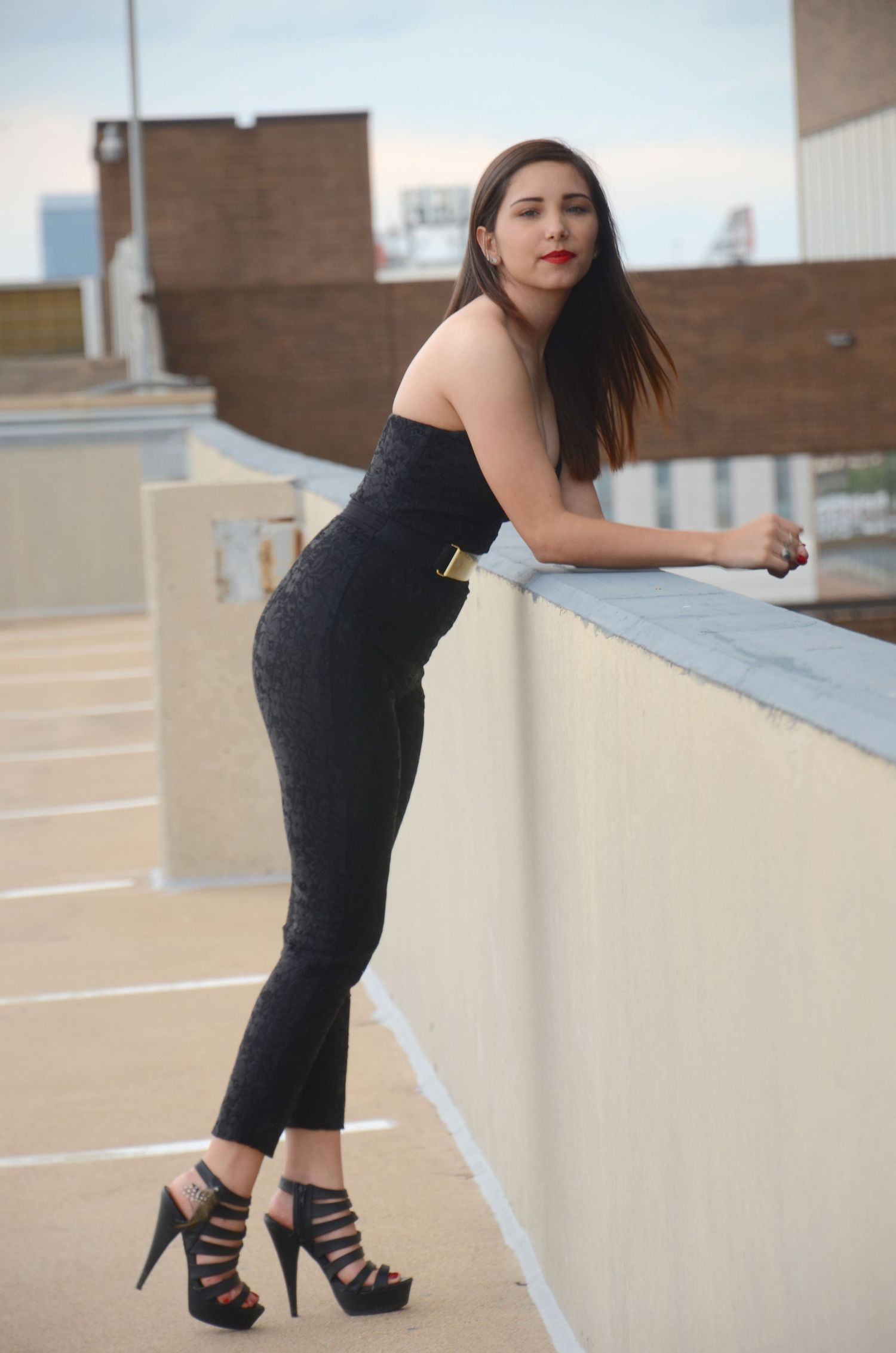 Fashionable woman in black jumpsuit poses on rooftop with high heels.