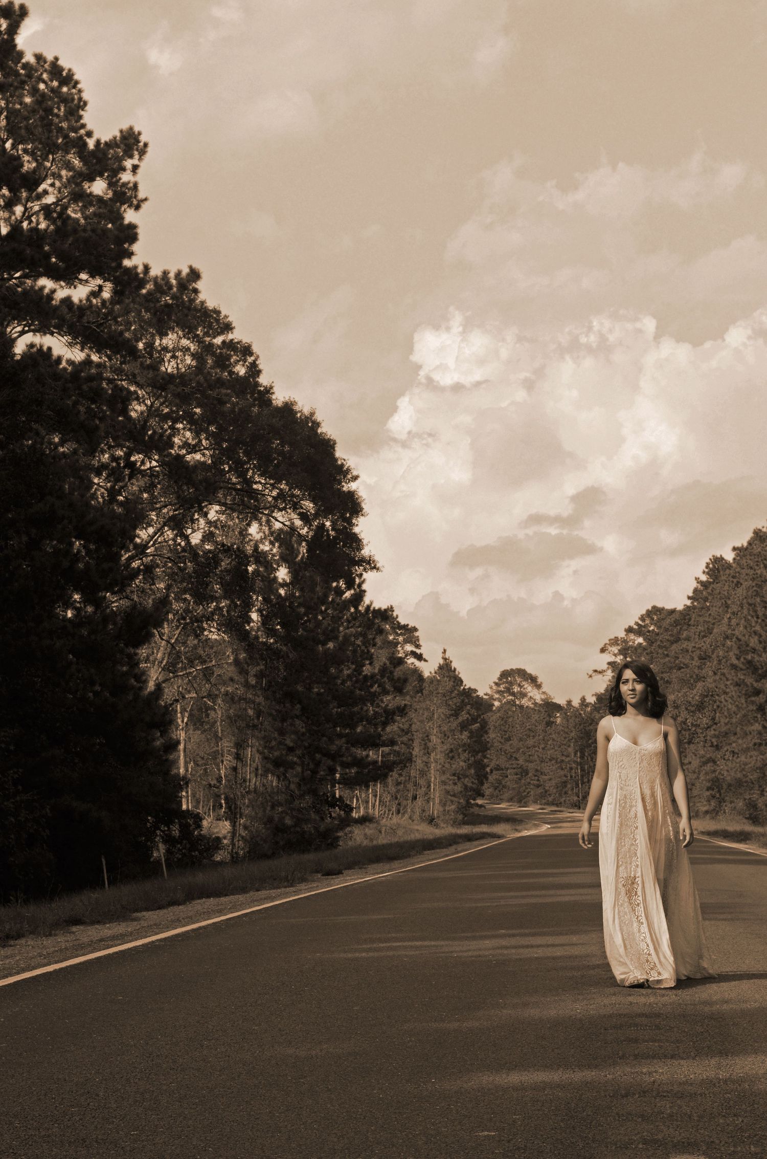 Person in flowing white dress stands on empty rural road surrounded by pine trees.