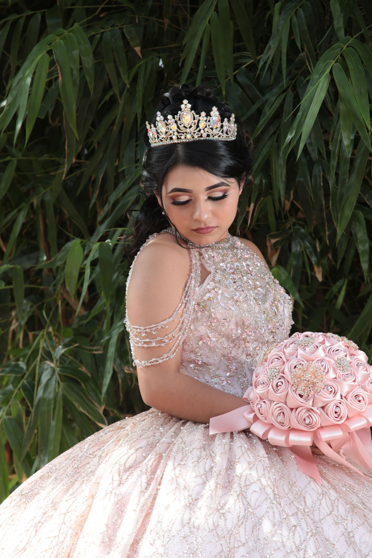 A close-up portrait showing a sparkly crown and pink gown against bamboo foliage.