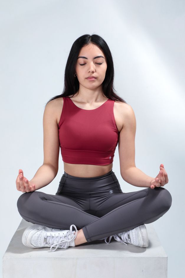 Woman in burgundy activewear meditating in lotus position on white background.