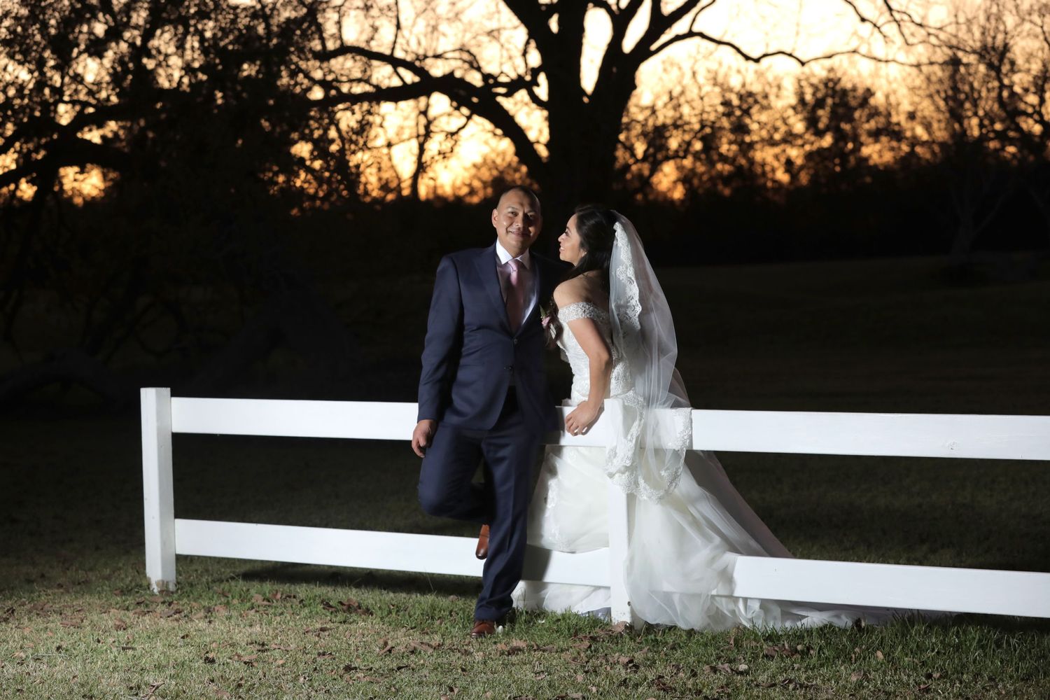 Couple in wedding attire poses by white fence at sunset.