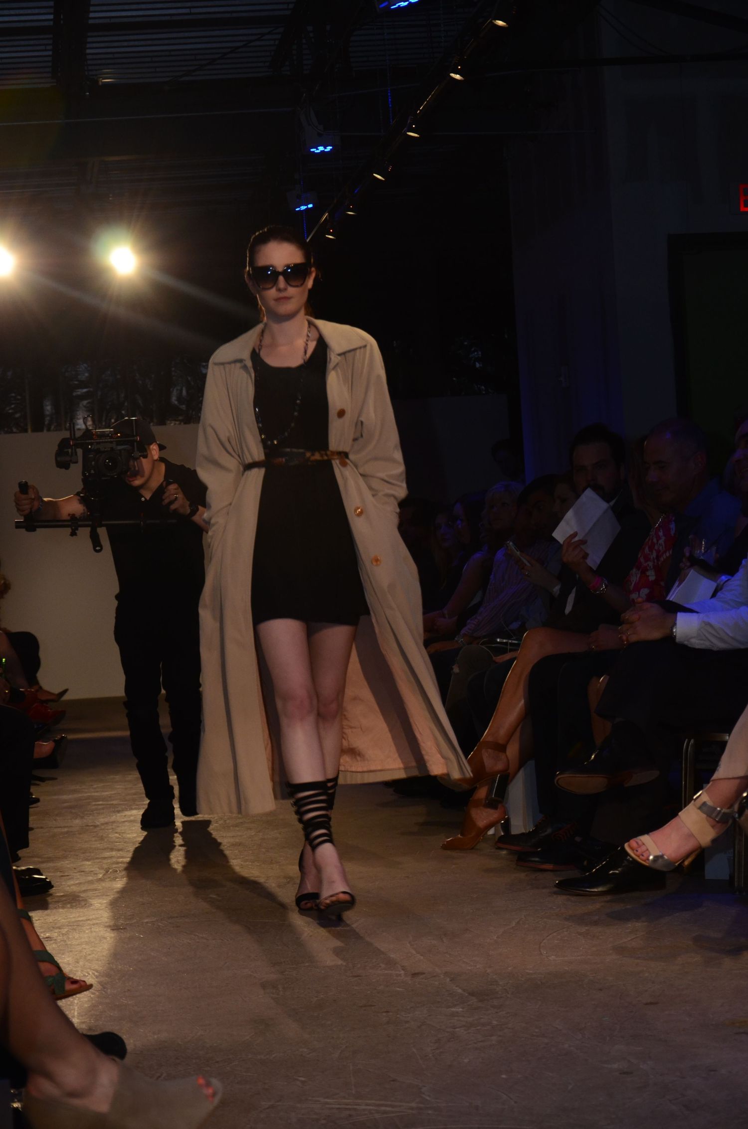Model walking runway in black dress and beige trench coat during fashion show.