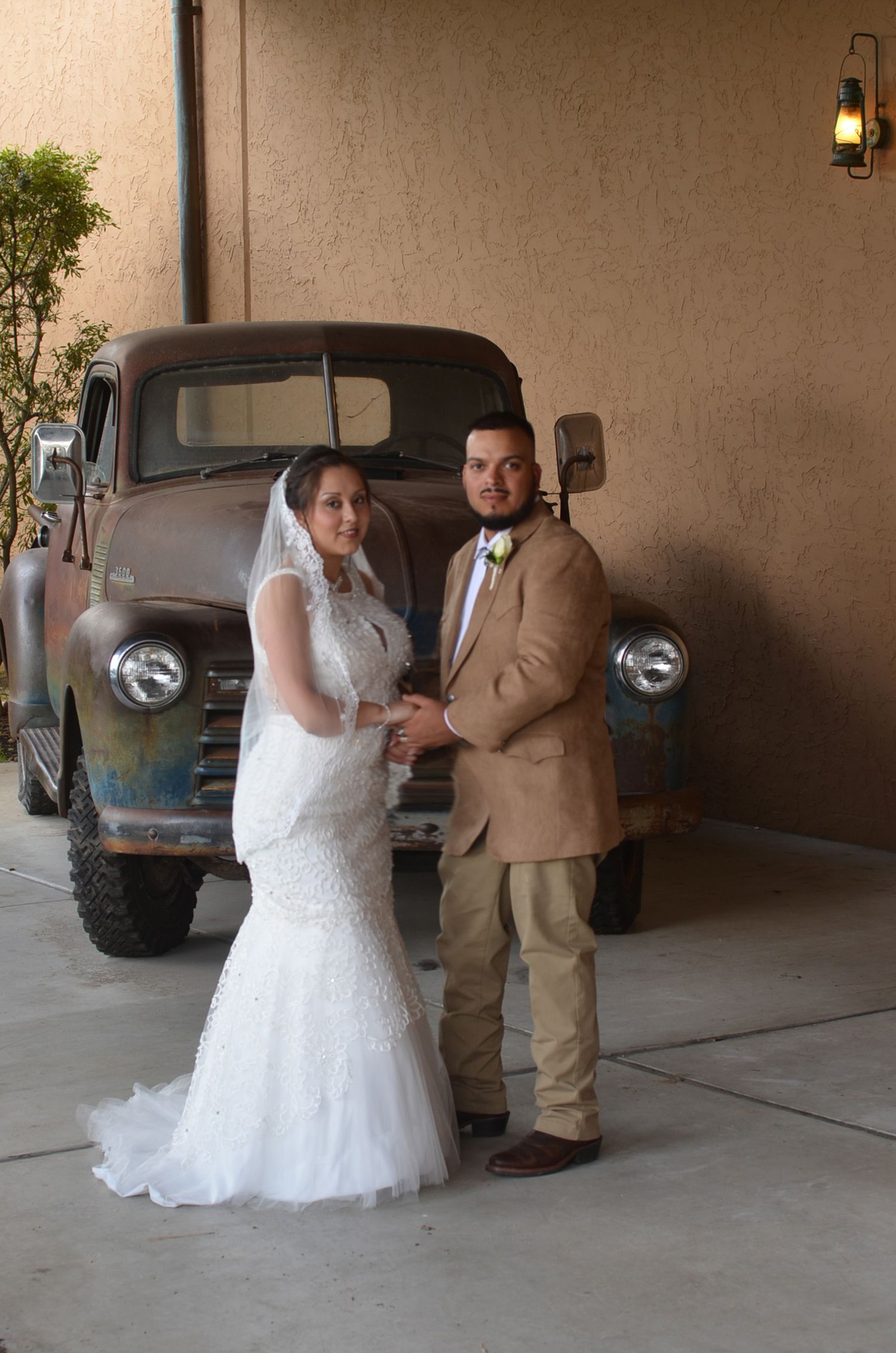 A couple in wedding attire poses in front of a classic blue pickup truck against a neutral wall.