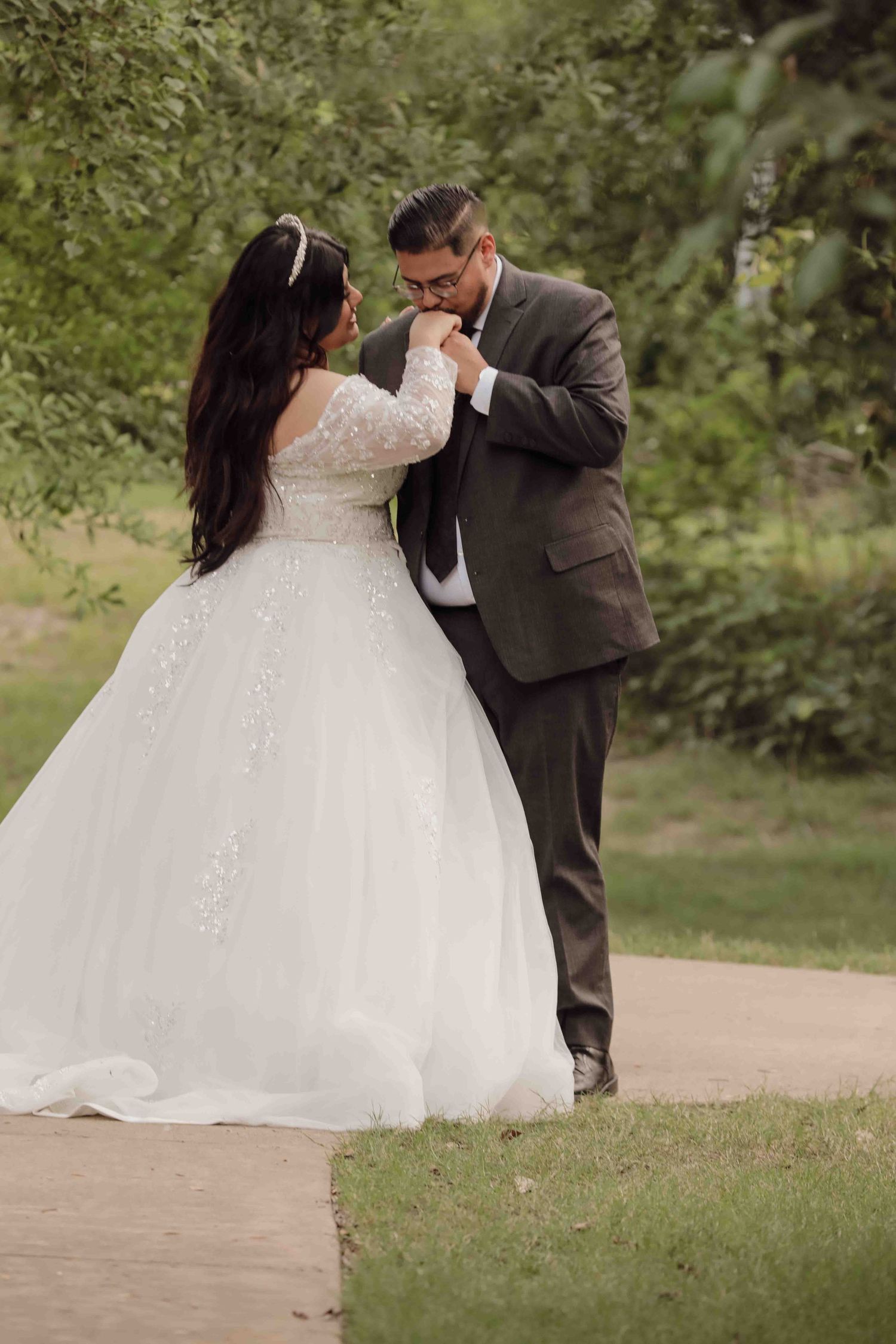 A romantic wedding moment captured outdoors with a bride in a lace sleeve ballgown and groom in grey suit on a garden path.