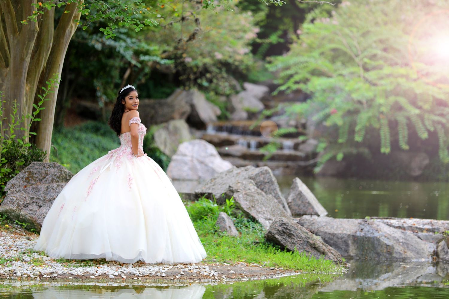 A person in a white ball gown poses in a dreamy garden setting near a pond.