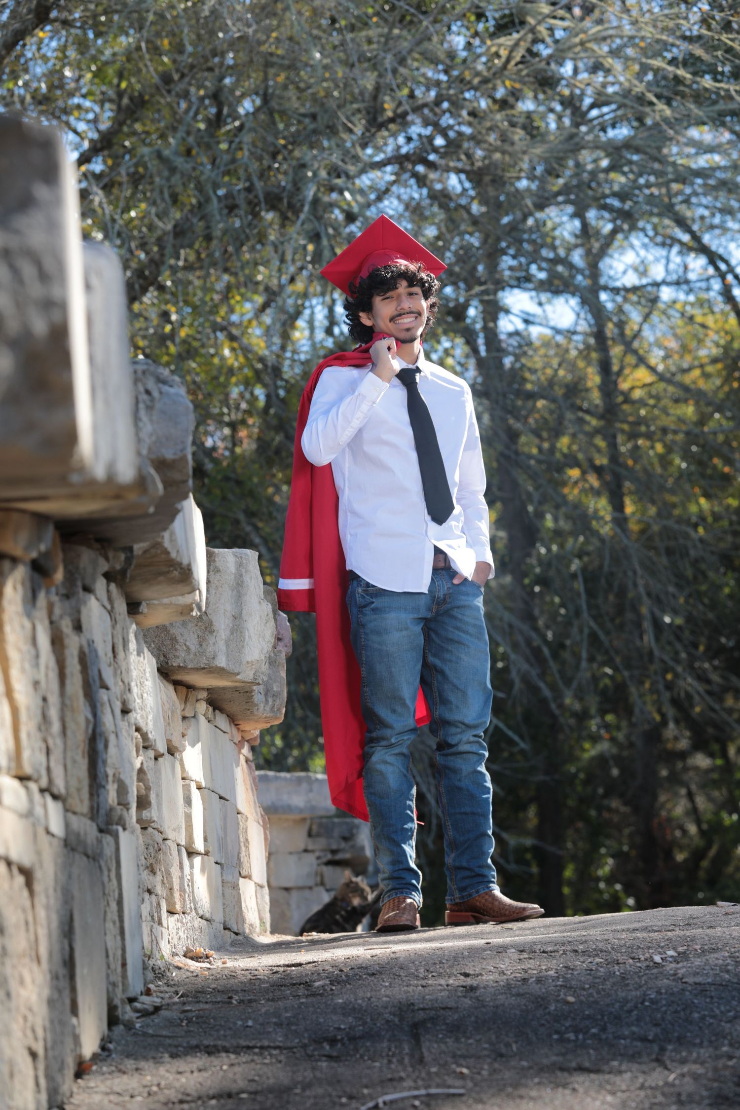 Graduate in red cap and gown standing on stone wall pathway.