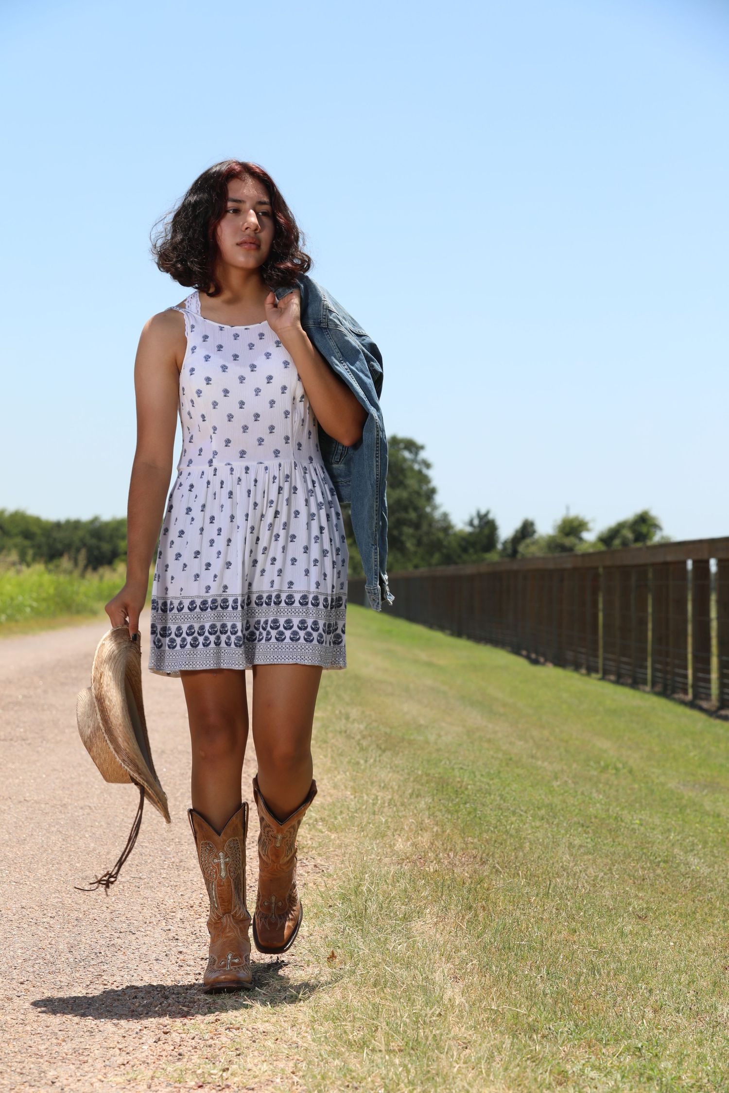 Woman in white summer dress and denim jacket walking along rural path with cowboy boots.
