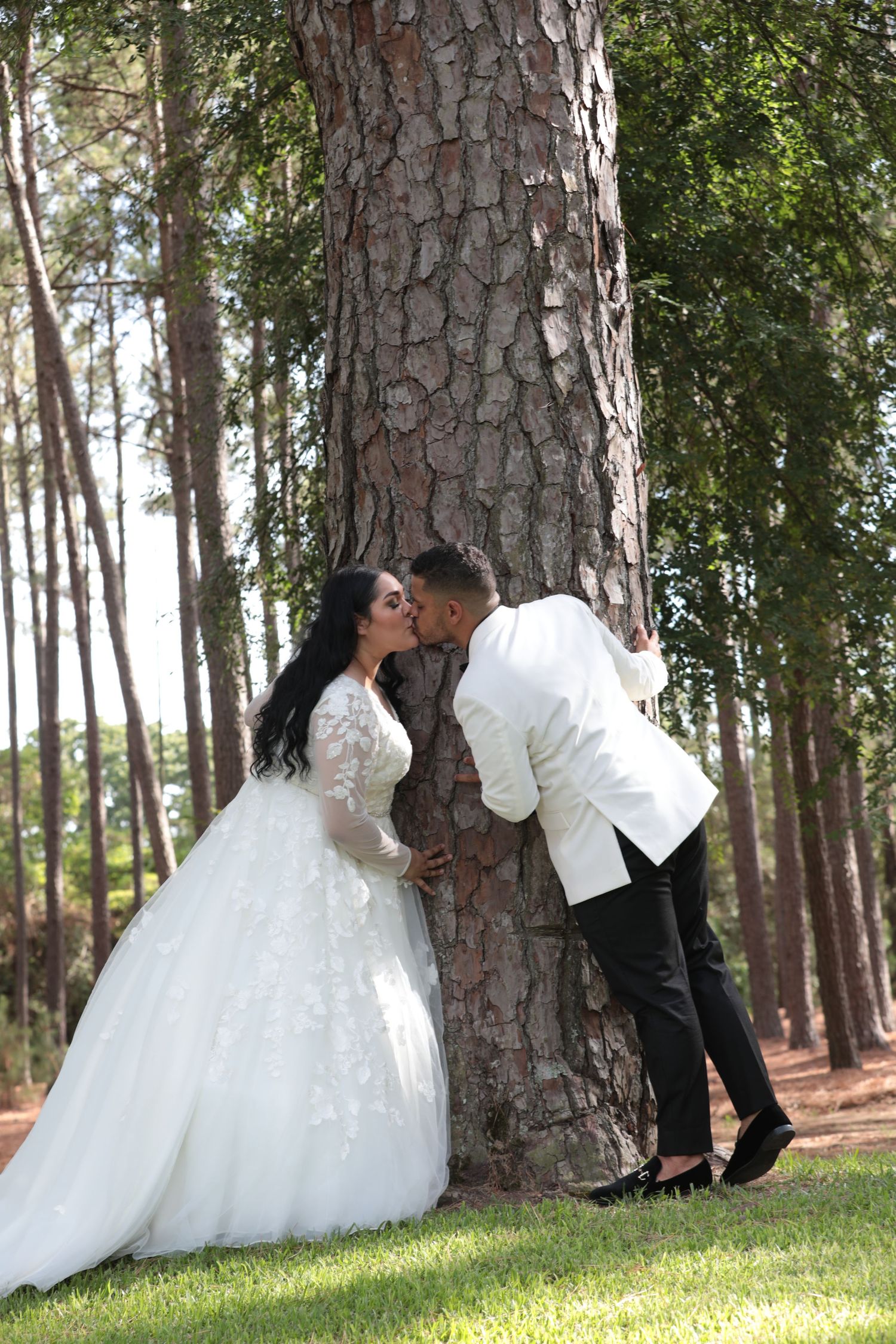 Wedding couple shares romantic moment against large pine tree in wooded setting.