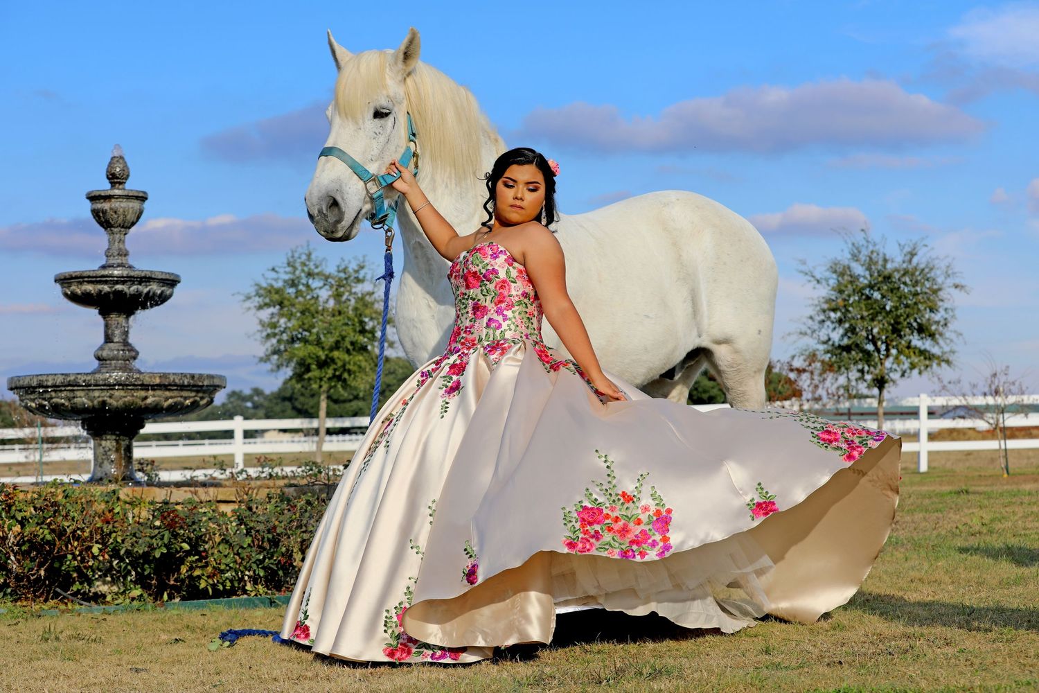A person in an embroidered white gown poses with a white horse near a fountain.