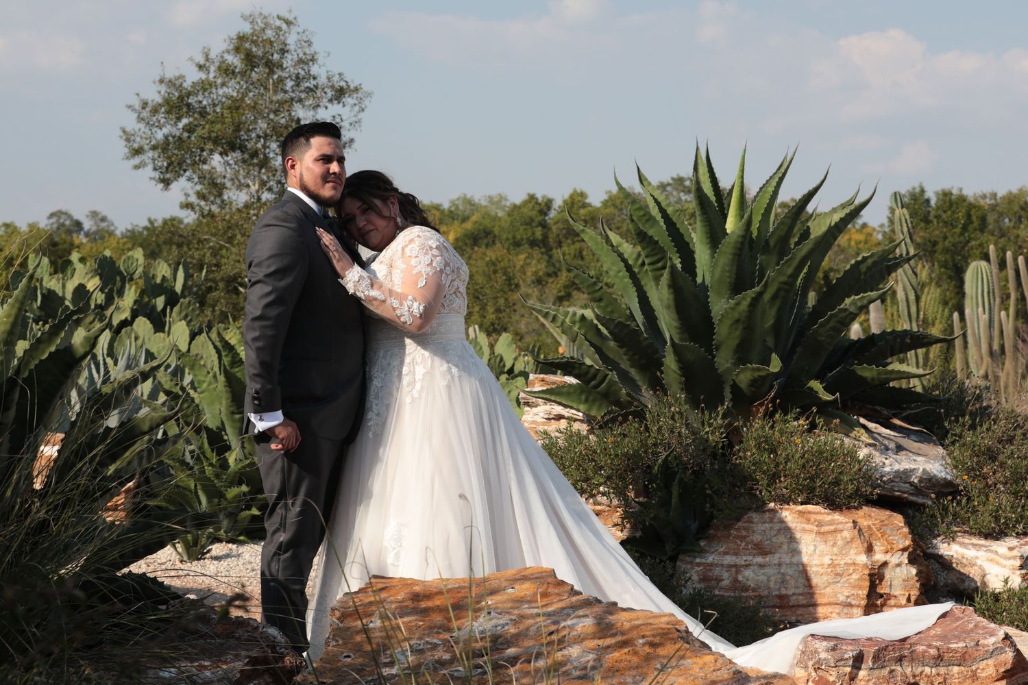 A romantic wedding couple embraces in front of large desert plants and stone features.