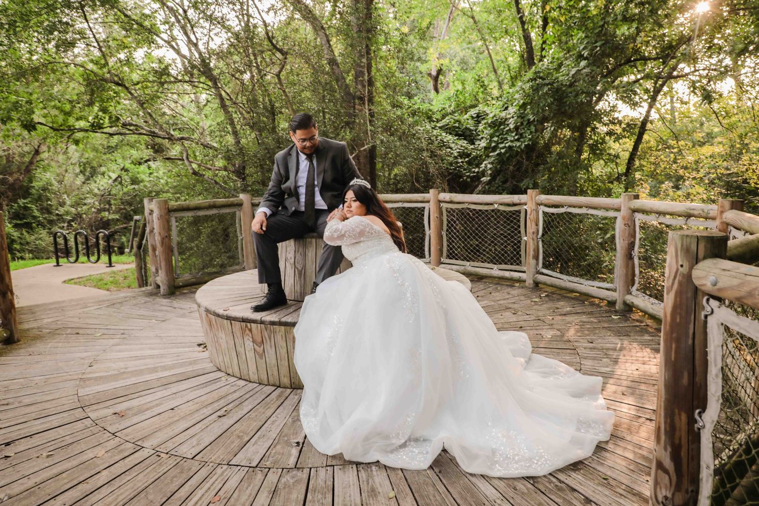 Karina&Eric newlywed couple shares a romantic moment on a wooden bridge pathway surrounded by lush greenery in a forest setting.