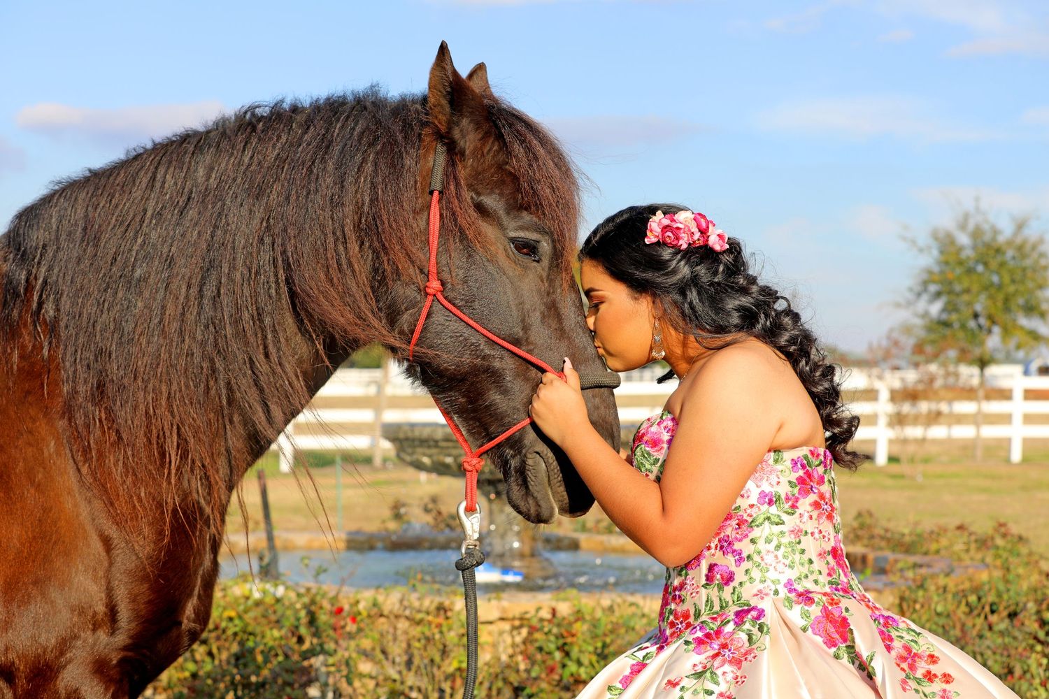 A brown horse and someone in a pink floral dress share a tender moment in a rural setting against a white fence.