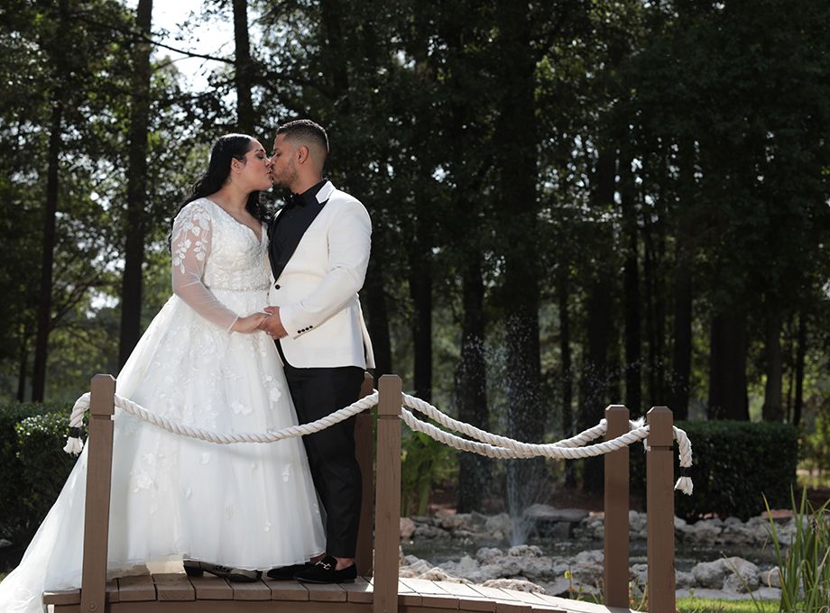 A couple shares a romantic moment on a wooden bridge in wedding attire surrounded by pine trees.