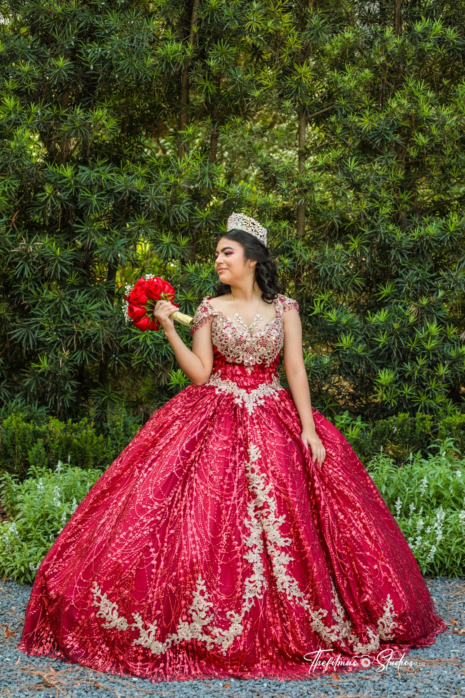 Elegant red quinceañera ball gown with gold embroidery details photographed in garden setting.