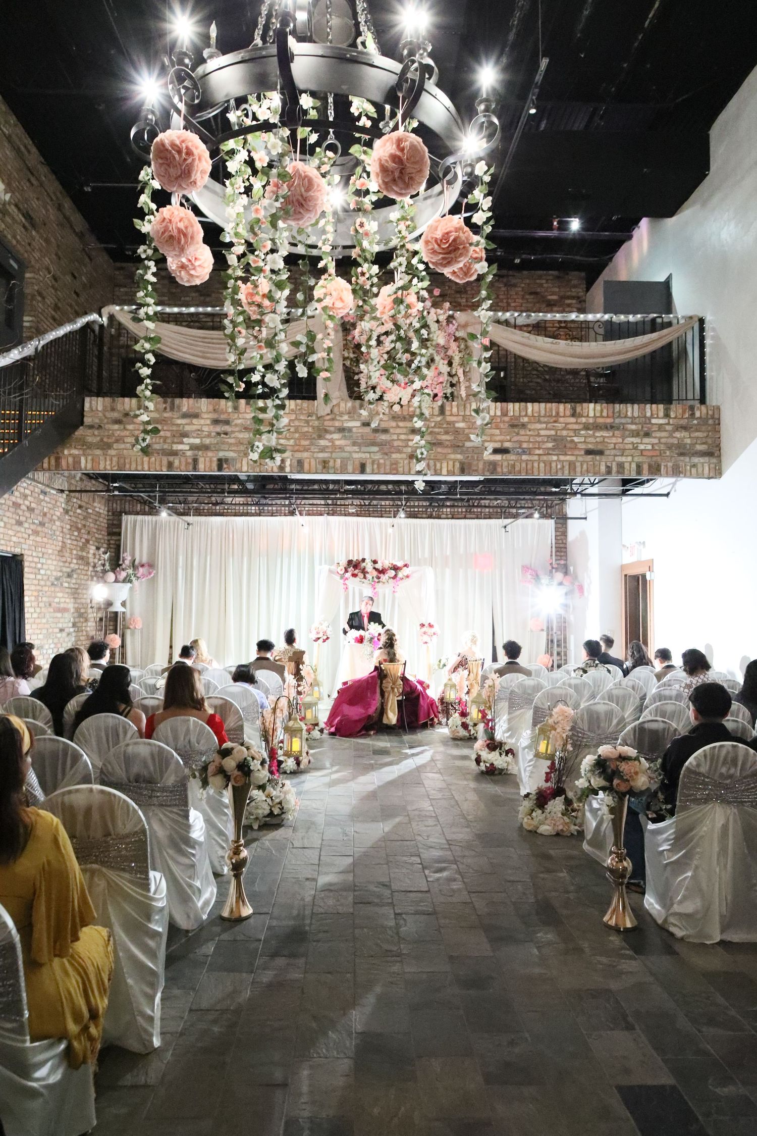 Elegant wedding ceremony setup with pink floral chandelier, white draping, and covered chairs in an industrial-style venue.