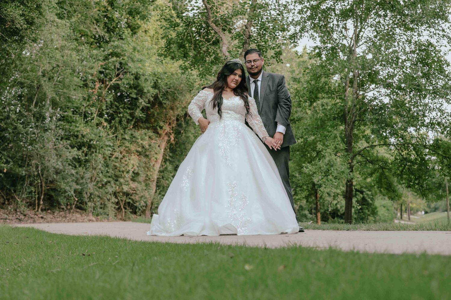 A romantic wedding couple stands together on a path surrounded by lush green trees and foliage.