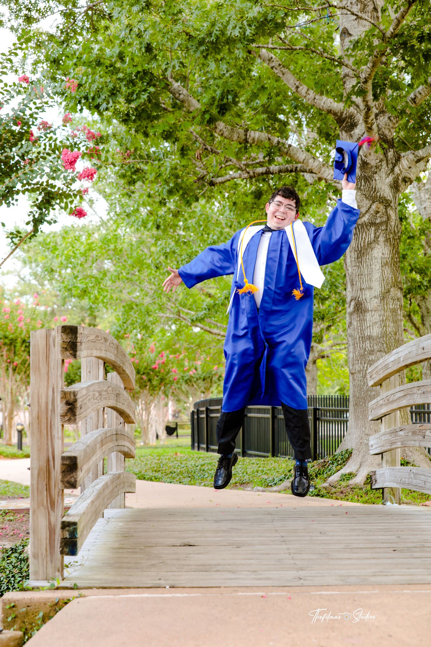 Graduate in blue cap and gown jumps joyfully on a wooden bridge surrounded by trees.