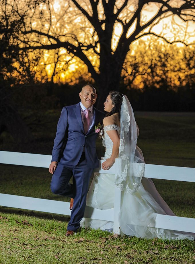 A newlywed couple stands by a white fence at sunset during their wedding photoshoot.