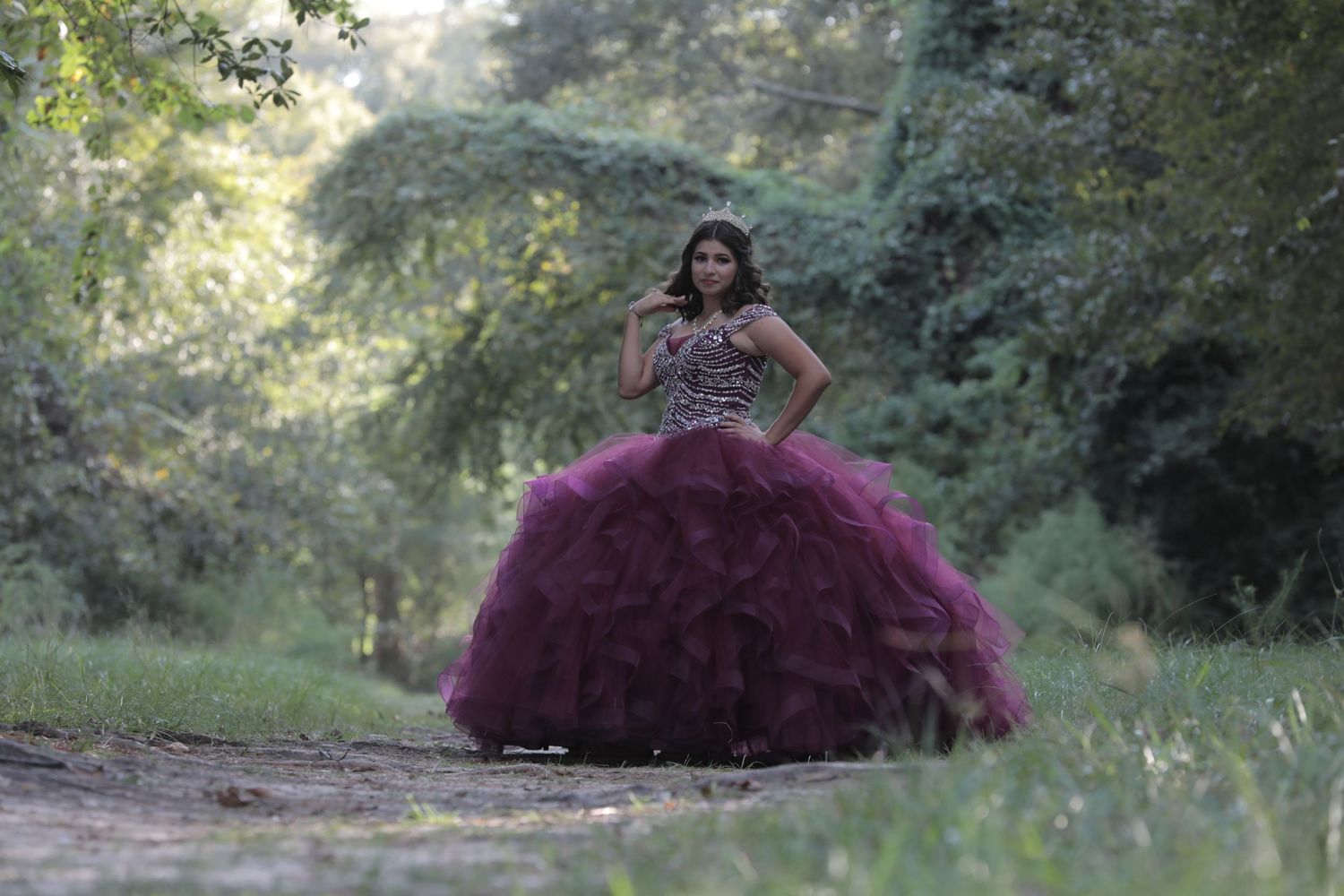 A person wearing a dramatic purple ball gown poses in a wooded outdoor setting.