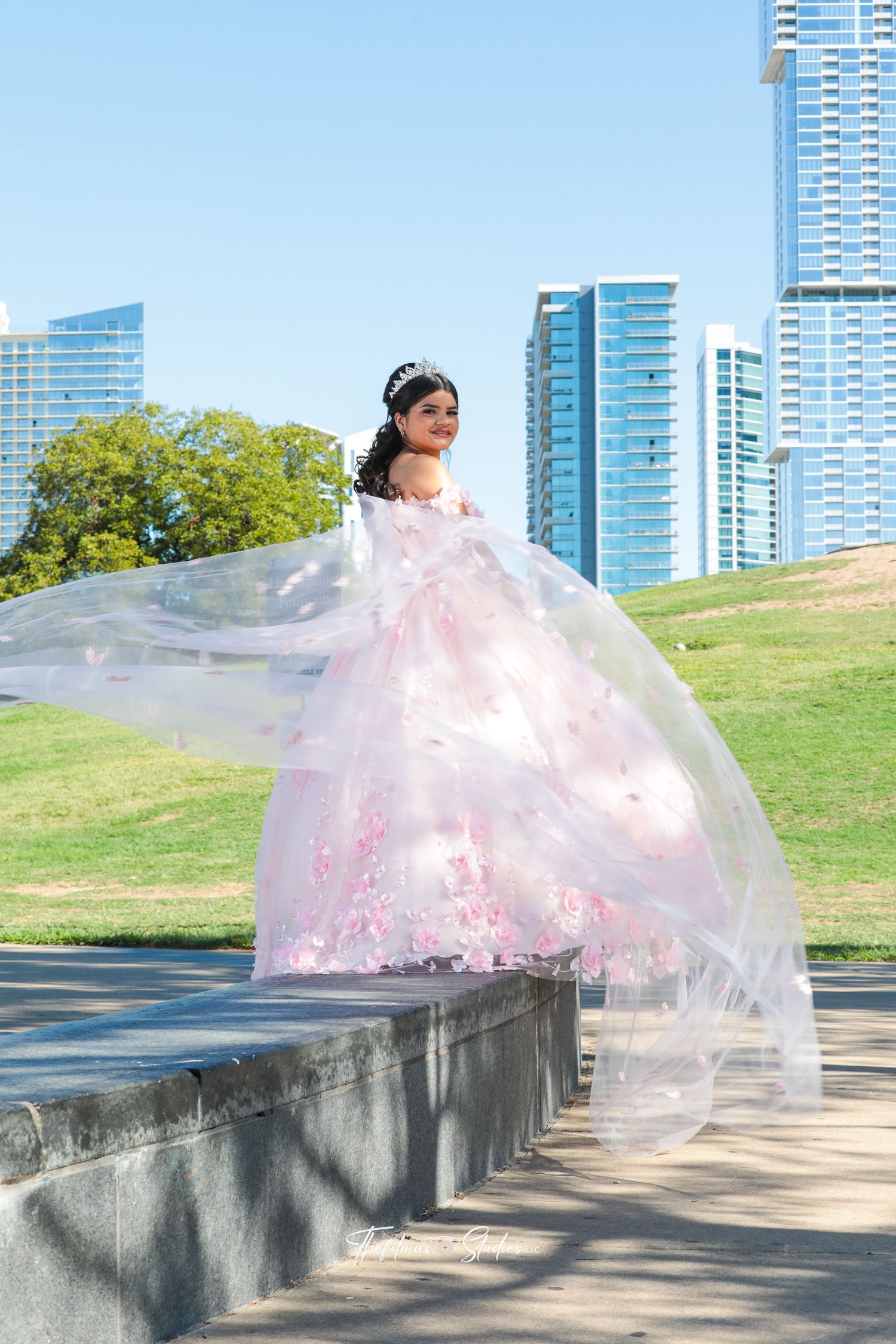 A pink quinceañera dress flows elegantly against a backdrop of modern city skyscrapers and green lawn.
