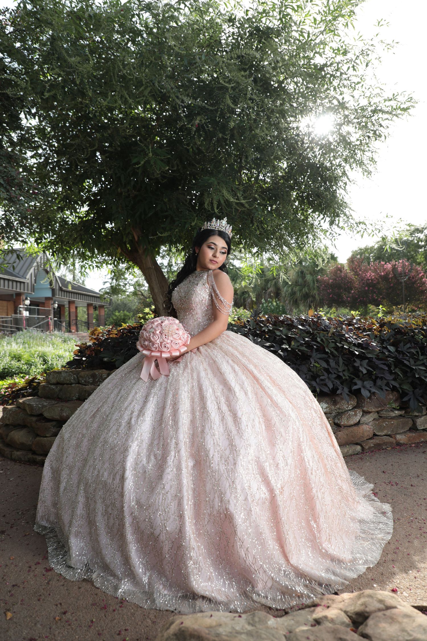 A person in a glittering pink ball gown poses near a tree with a pink bouquet.