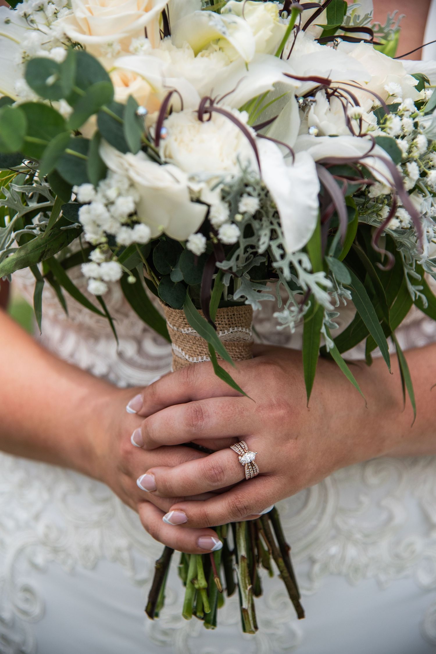 Close-up detail of hands holding a white floral wedding bouquet with an engagement ring and greenery.