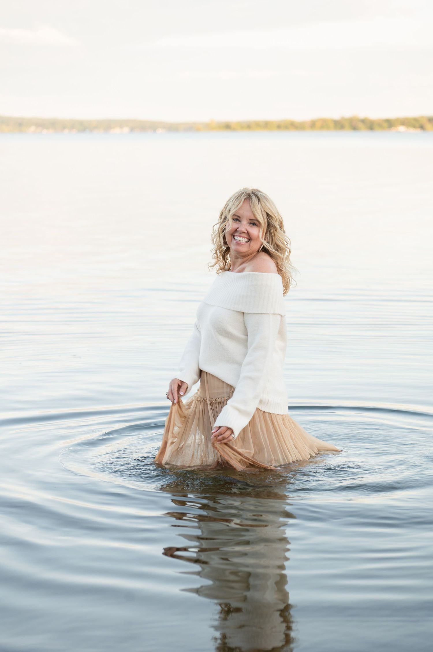 Woman wearing white sweater and beige skirt standing in tranquil lake water.