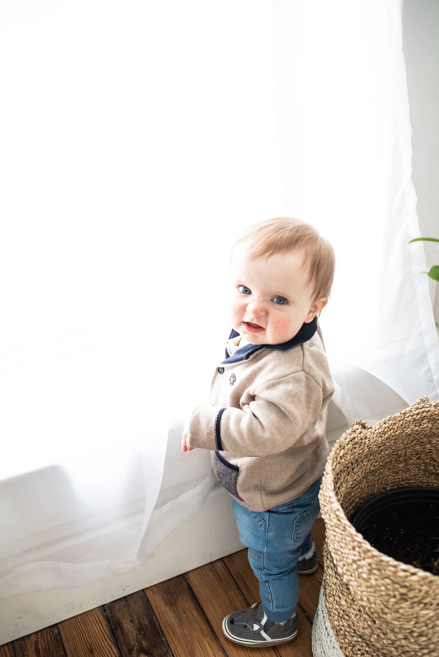 Toddler stands by a woven basket near a bright window.